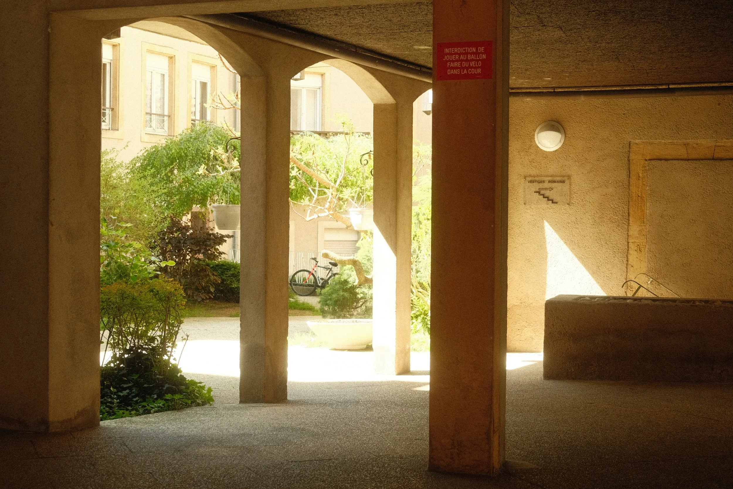 Vue d'un passage couvert menant à un jardin avec des plantes, un vélo et une statue. Murs en pierre avec deux panneaux, l'un rouge avec une inscription en français.
