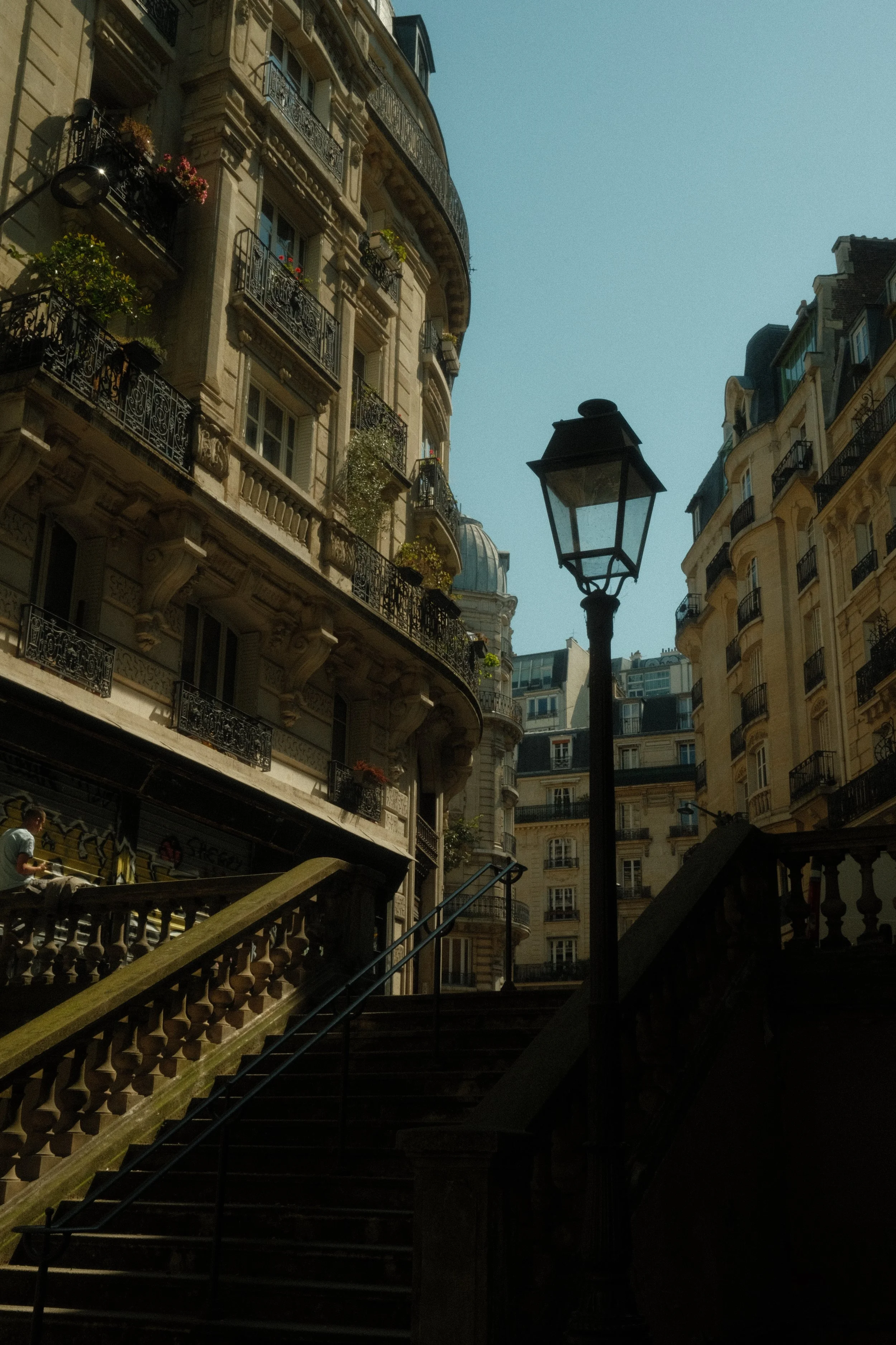 Escalier menant à une rue pavée bordée d'immeubles parisiens haussmanniens avec balcons en fer forgé, un lampadaire en fer noir au centre et un ciel bleu clair.
