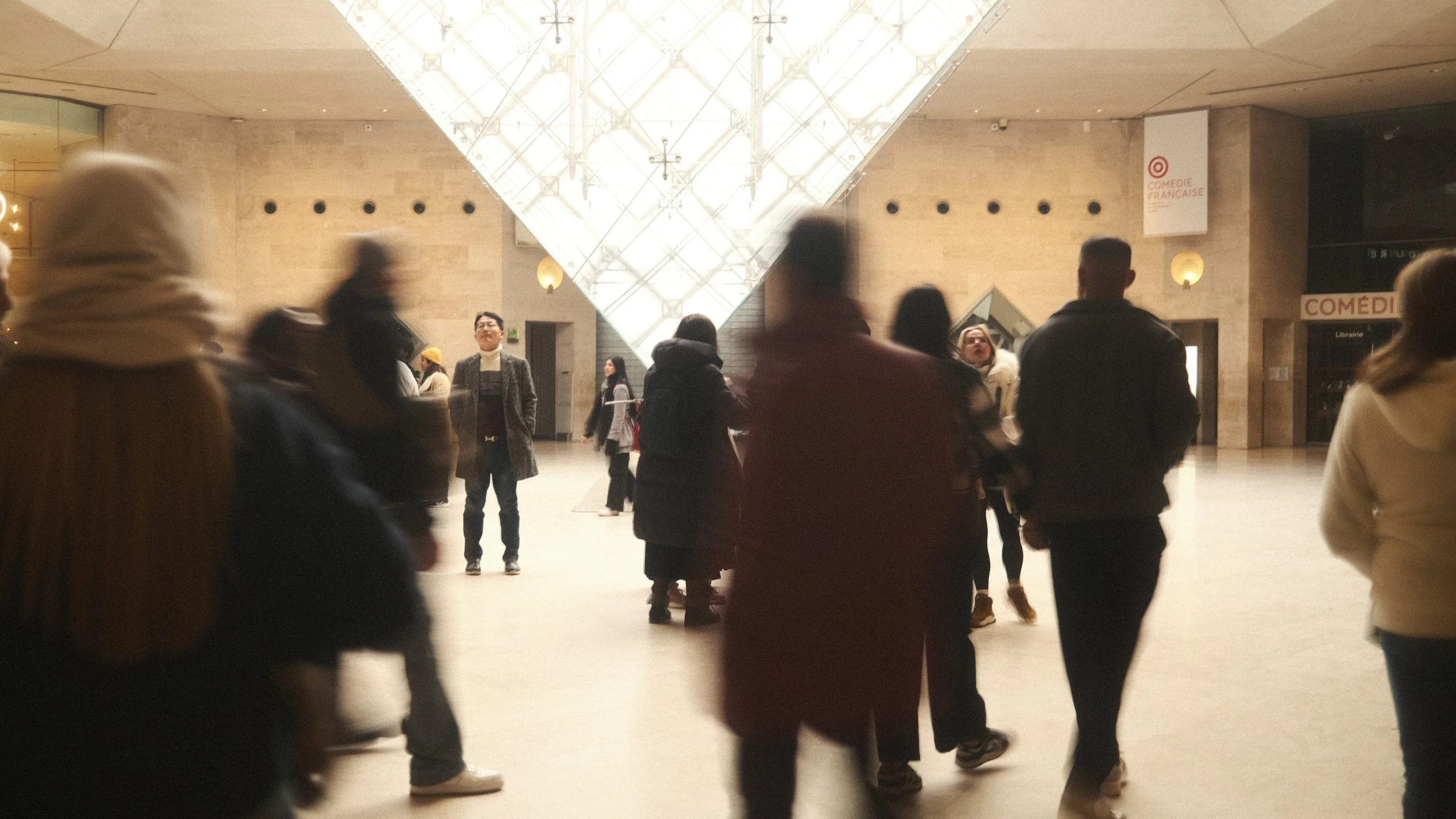 Une foule de personnes dans un hall spacieux avec un plafond en verre triangulaire lumineux, murs en pierre, et une boutique librairie à droite.