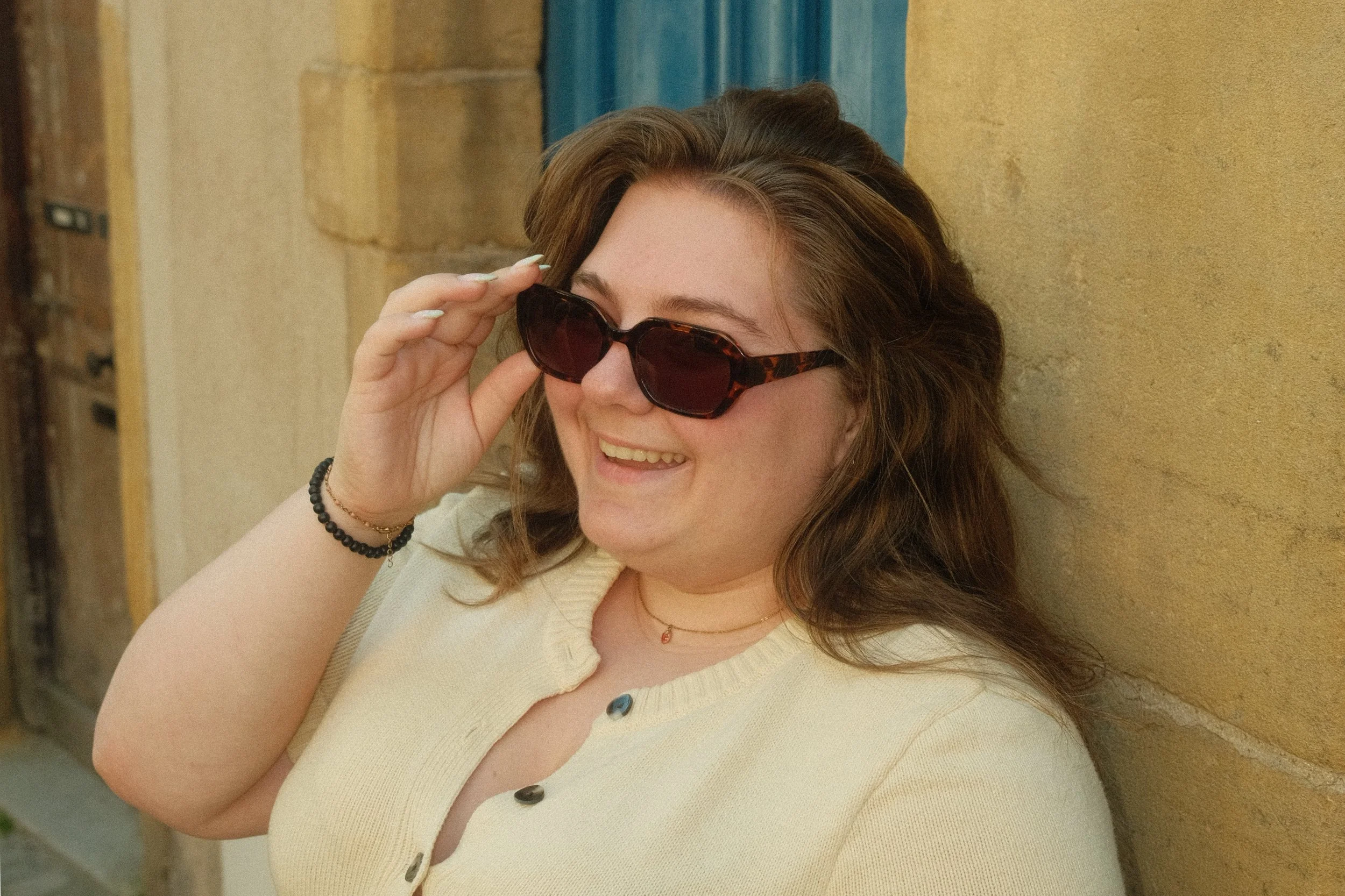 Une femme souriante portant des lunettes de soleil, vêtue d'un haut jaune avec des boutons, se tient contre un mur beige.