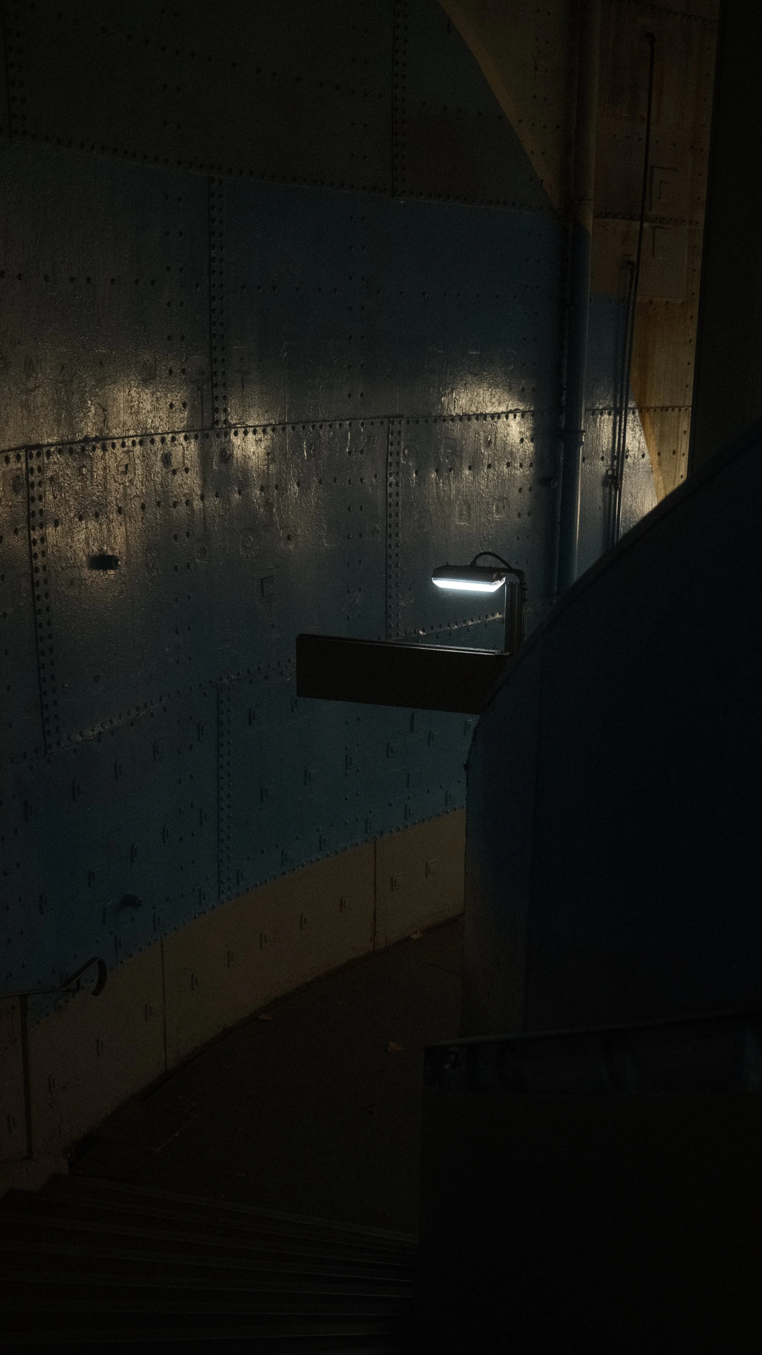 Escalier dans une zone sombre avec un mur en métal riveté et une lampe suspendue.