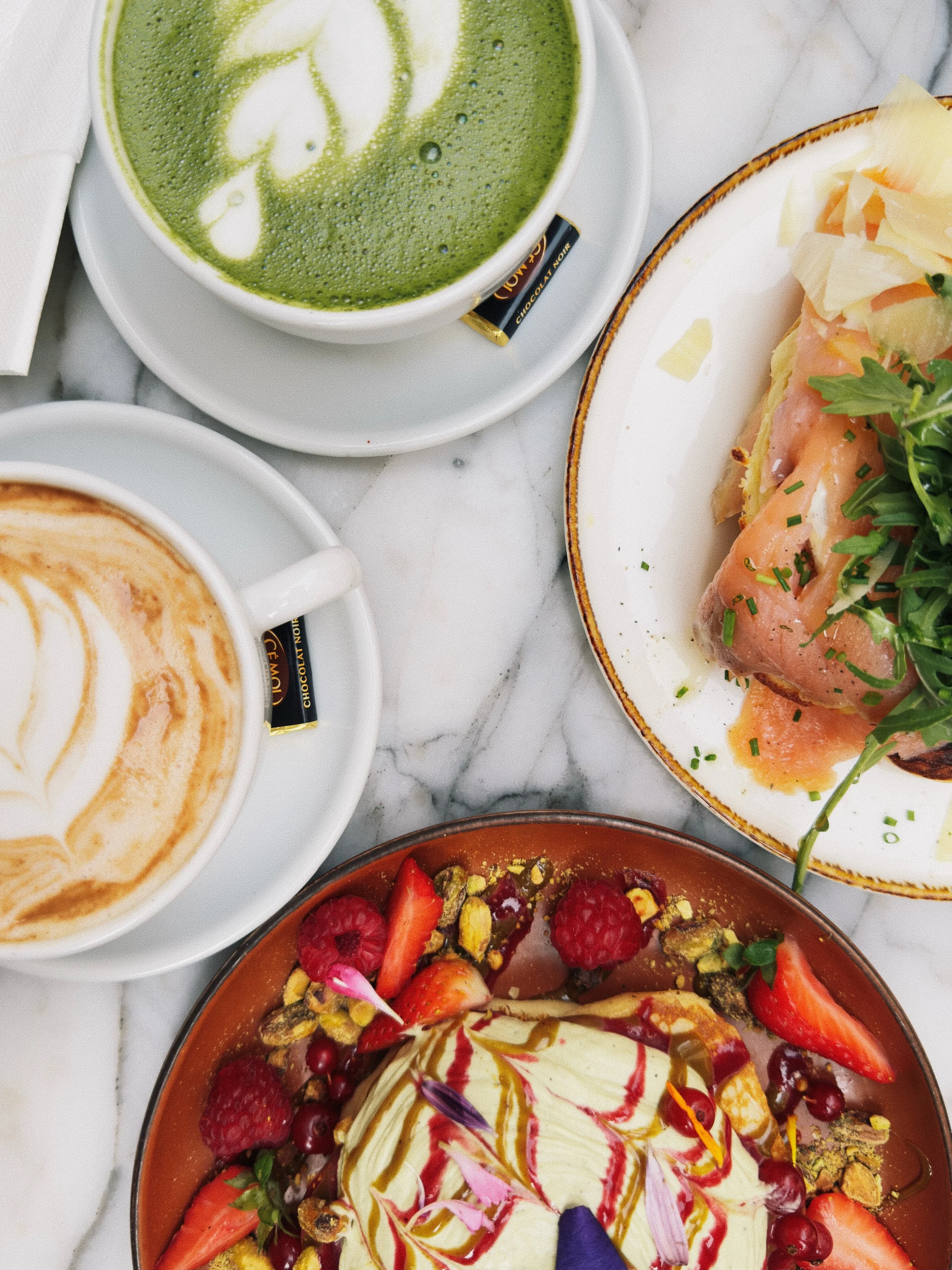Deux tasses de café avec de la crème en forme de cœur, une assiette de sandwich avec du jambon et des légumes, et un bol de dessert avec des fruits, des pistaches et une crème. La table est en marbre blanc.