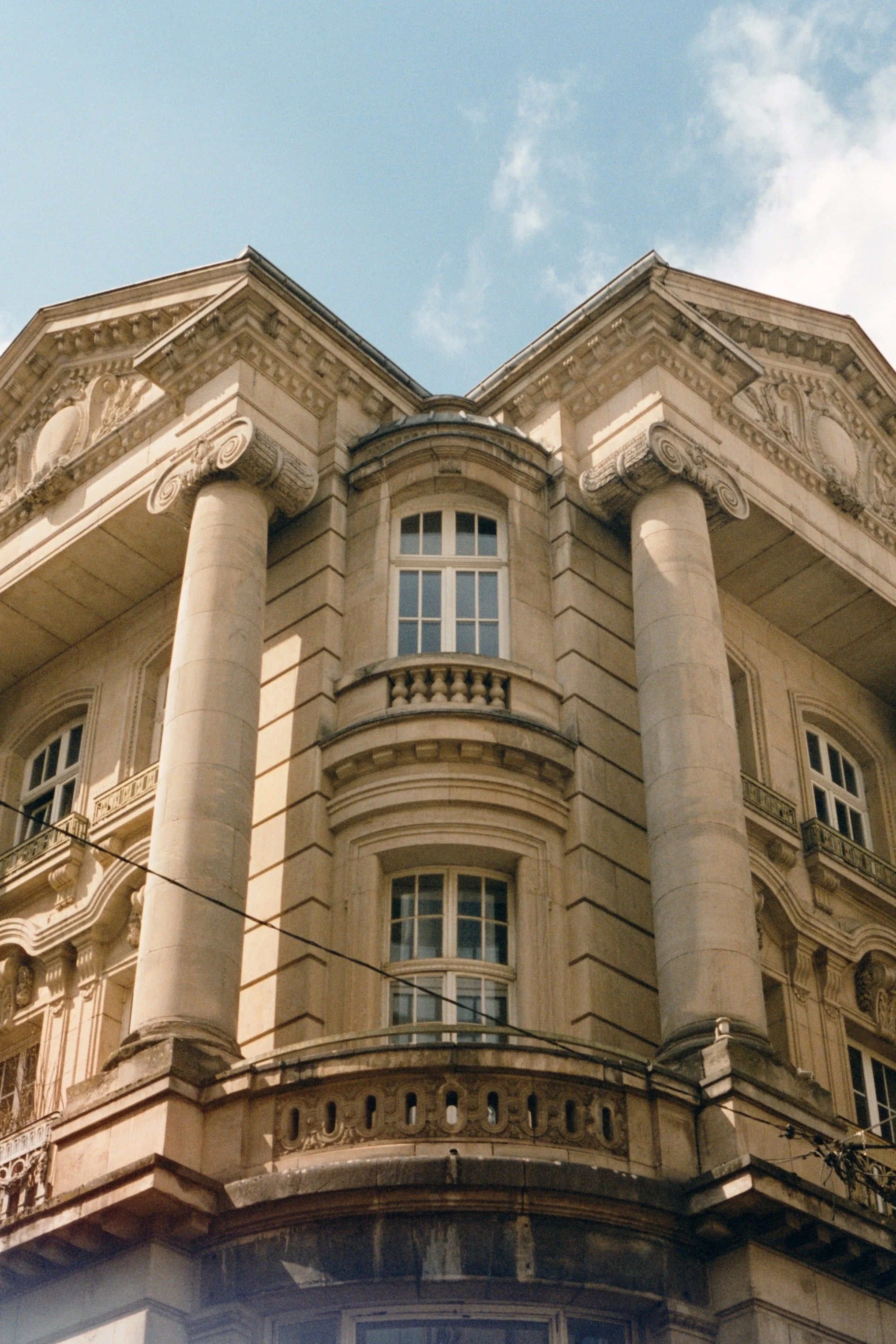 Façade d'un bâtiment historique en pierre avec des colonnes corinthiennes et de grandes fenêtres, sous un ciel bleu avec quelques nuages.