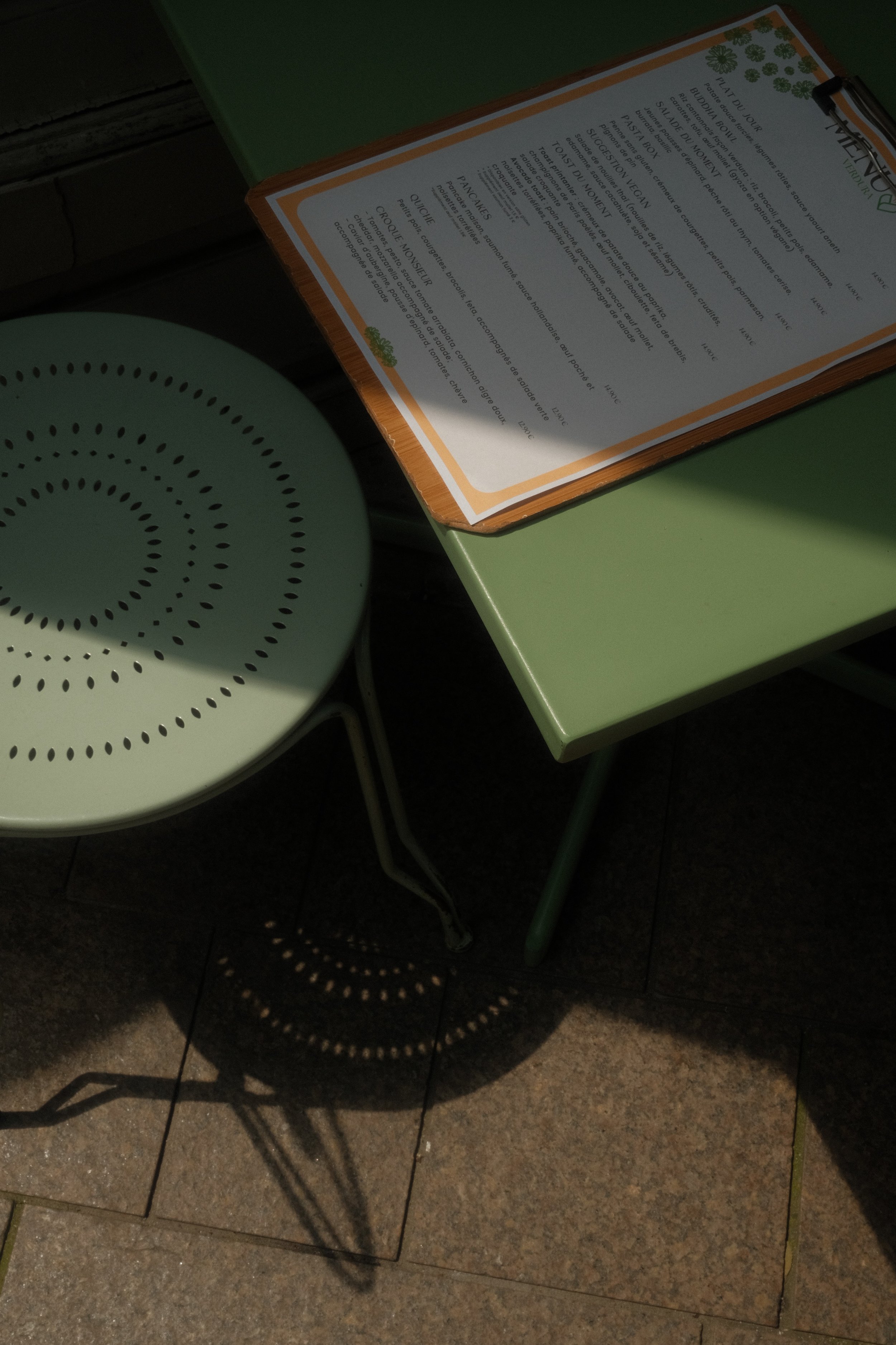 Une table vert clair avec un menu posé dessus et une chaise en métal vert clair avec un trou en motifs circulaires, la lumière du soleil crée une ombre circulaire sur le sol en carreaux en dessous de la chaise.