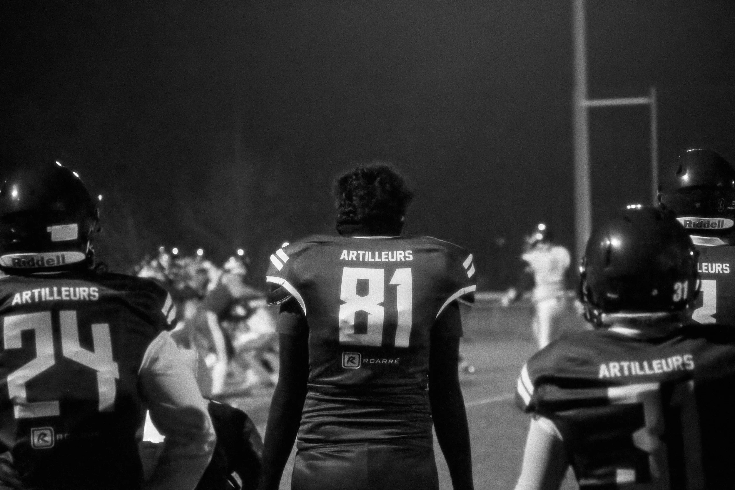 Groupe de joueurs de football américain lors d'un match, photographié de dos, portant des uniformes avec le mot "ARTILLEURS" et des numéros, sur un terrain nocturne.