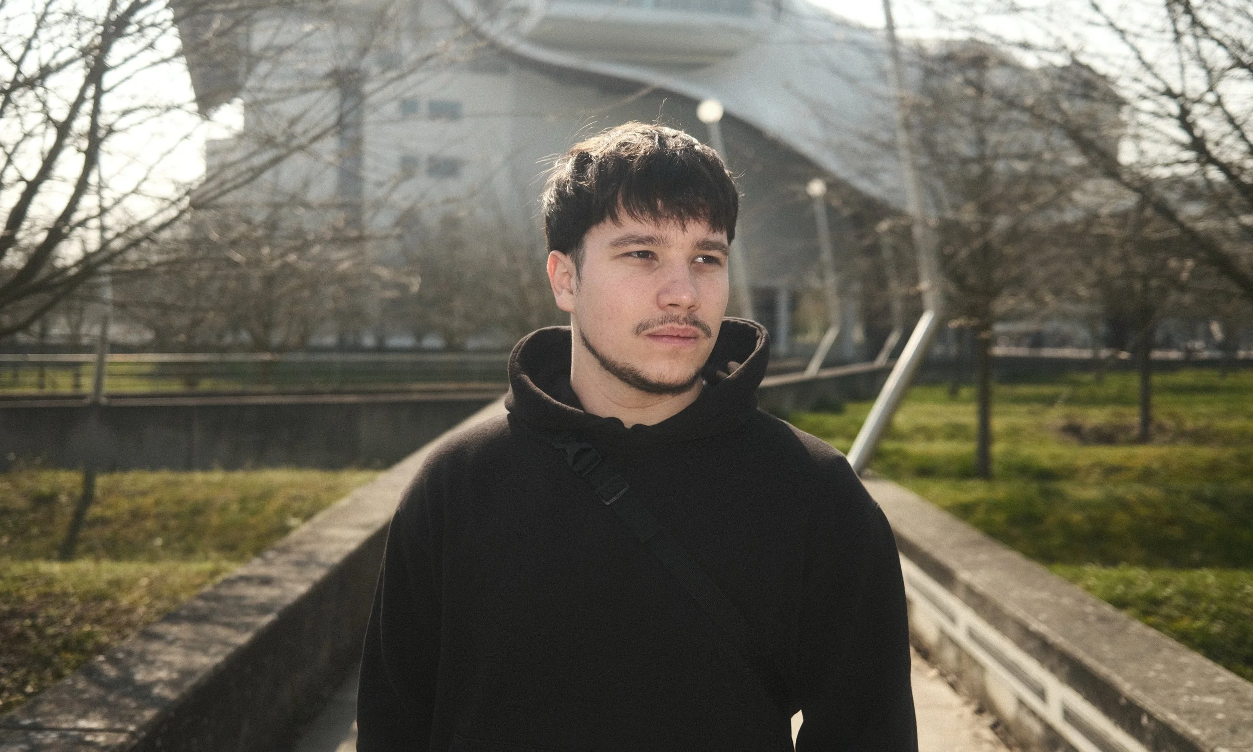 Jeune homme avec un sweat noir, portant une sacoche, marche dans un parc par une journée ensoleillée.