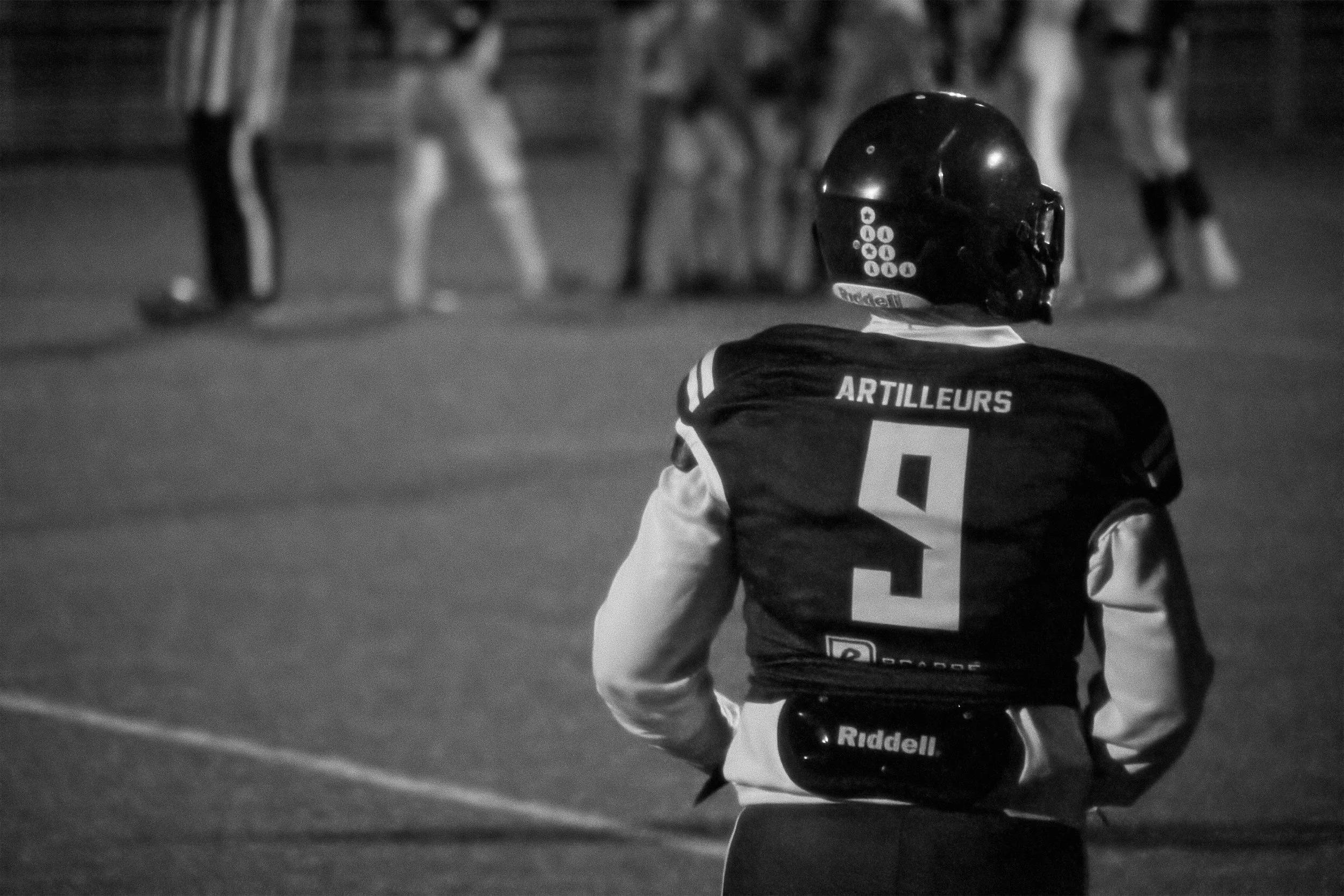 Un joueur de football américain portant un casque et un maillot avec le nom "ARTILLEURS" vu de dos, observant le terrain lors d'un match de nuit.