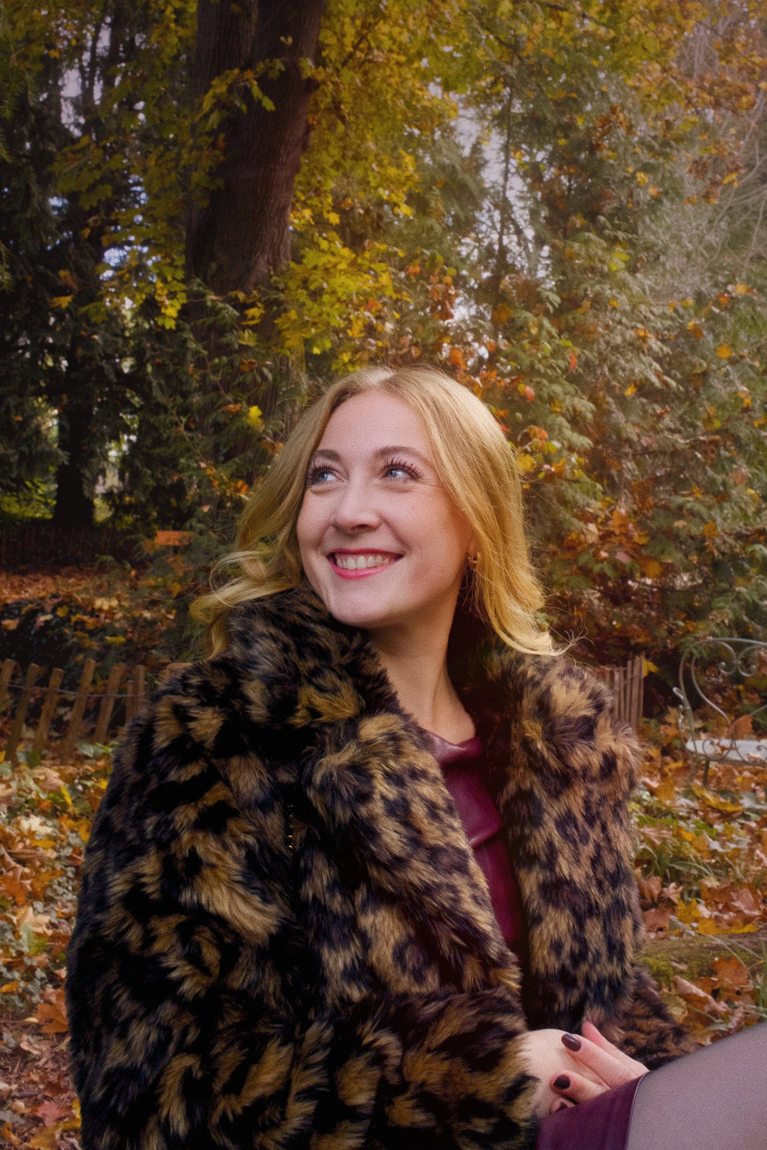 Jeune femme souriante avec une veste en fourrure à motif léopard, dans un parc en automne avec des feuilles tombées et des arbres colorés.