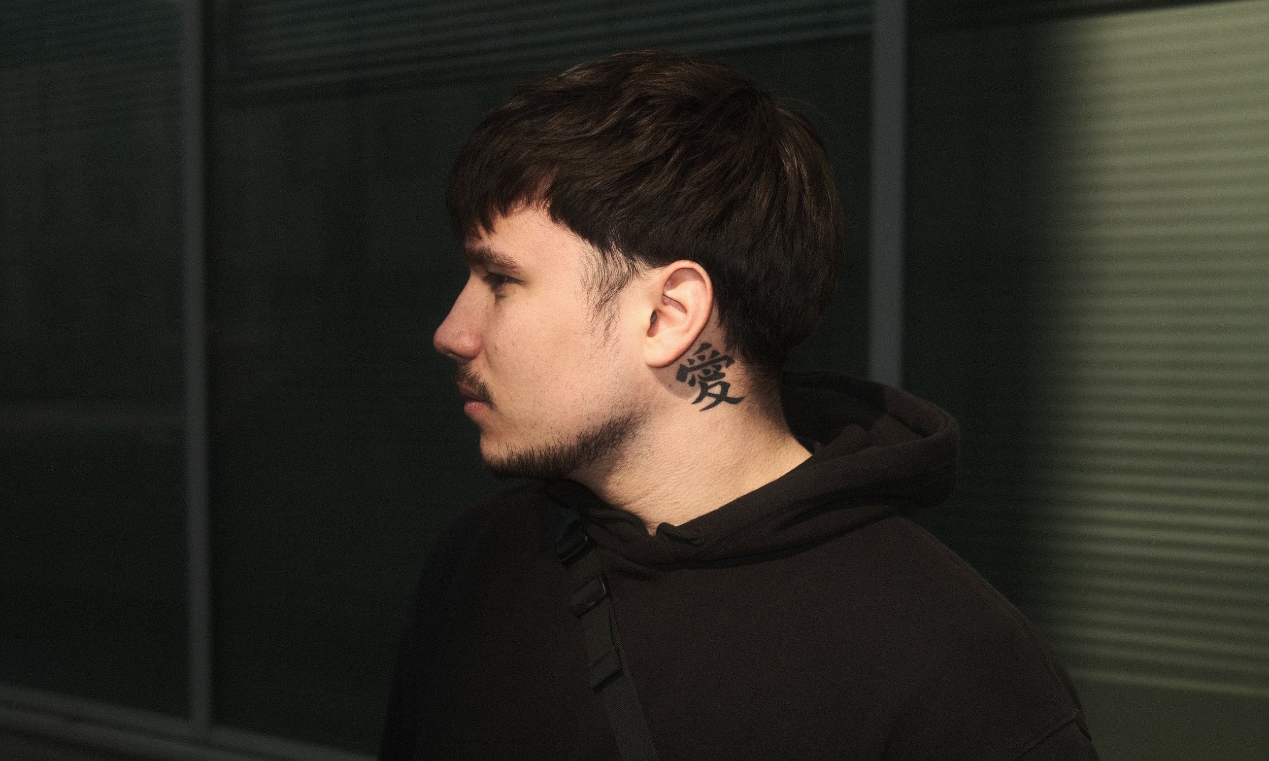 Jeune homme en profil, tatouage sur le cou, portant un sweat noir, dans un environnement sombre.