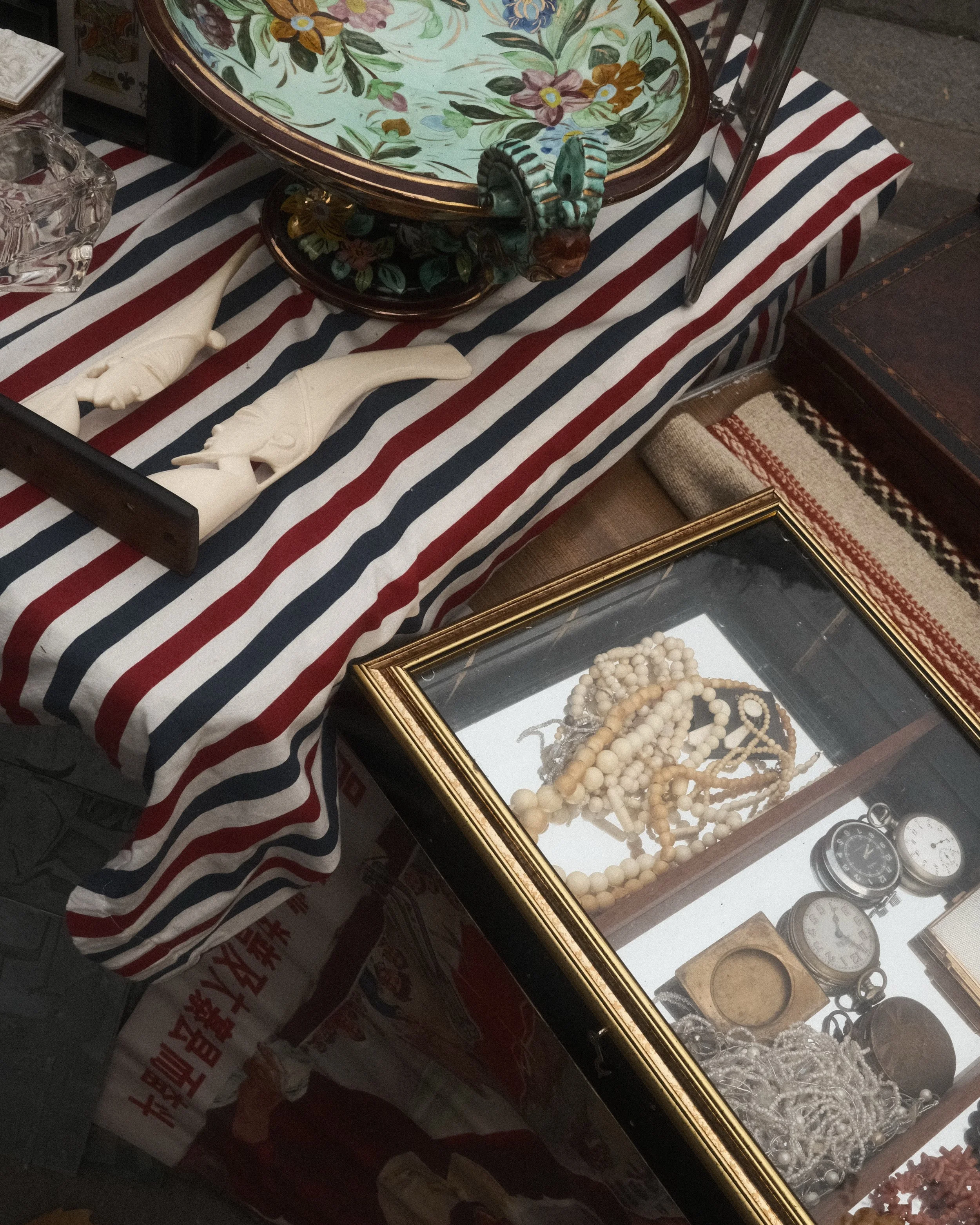 Une table recouverte d'une nappe à rayures rouges, blanches et bleues, avec des objets de collection tels que des bijoux, des montres anciennes, des figurines en os, une boîte en verre contenant des perles et d'autres petits objets, ainsi qu'un plate