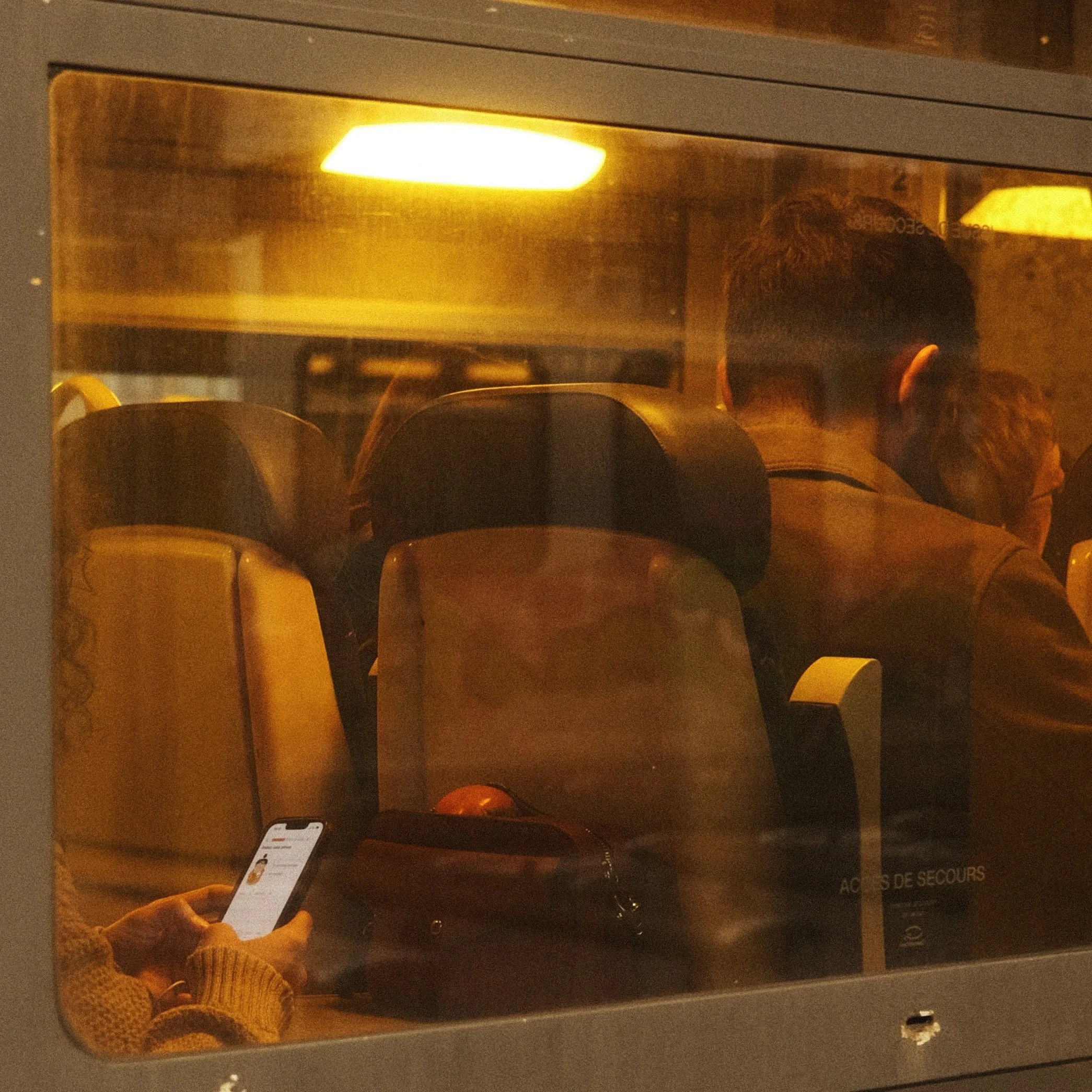 Vue à travers la vitre d'un train montrant plusieurs passagers assis, dont une personne regardant son téléphone portable, dans une ambiance chaleureuse et sombre