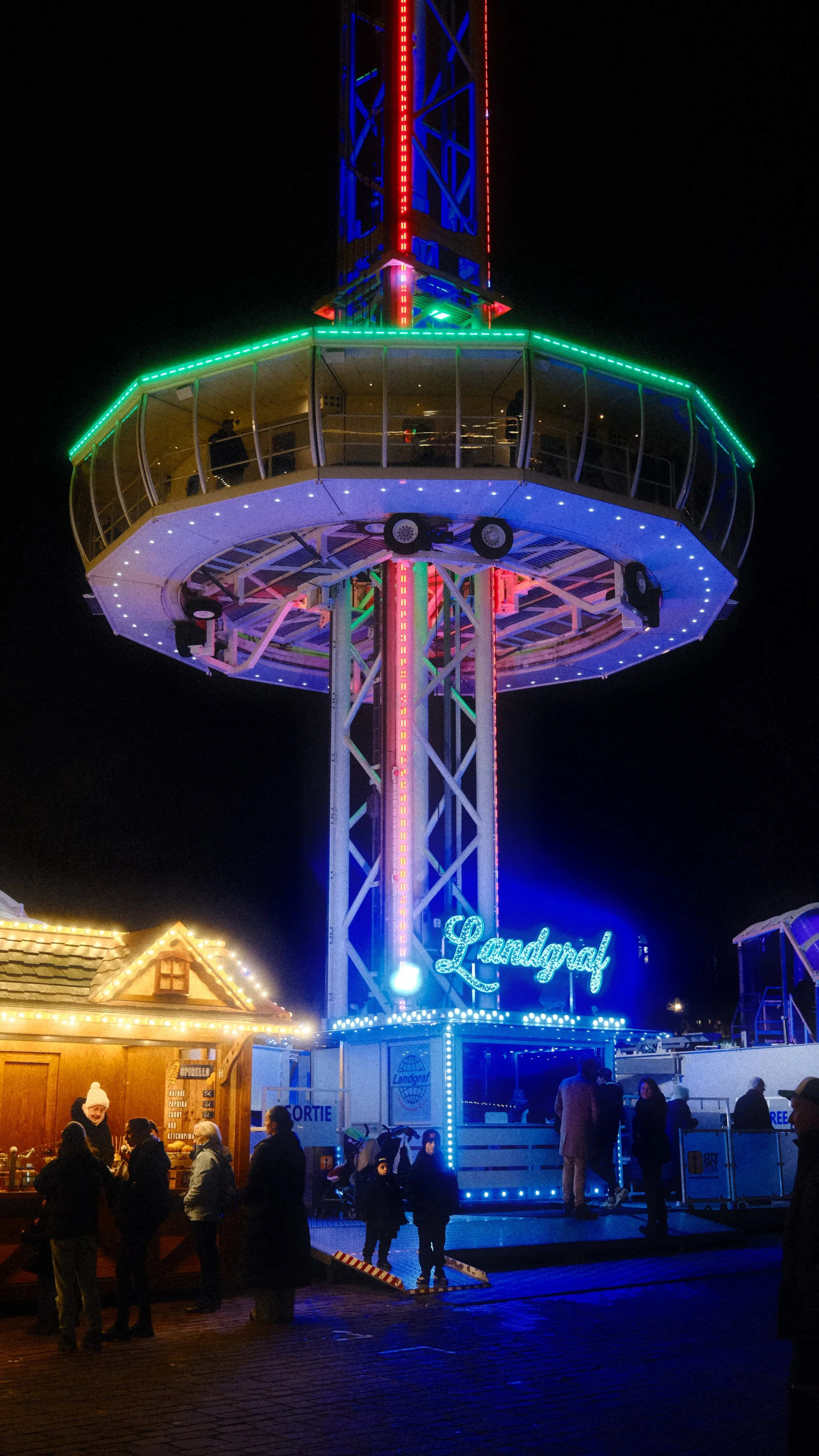 Une grande tour panoramique d'attraction illuminée de lumières colorées la nuit, avec des personnes faisant la queue en dessous.