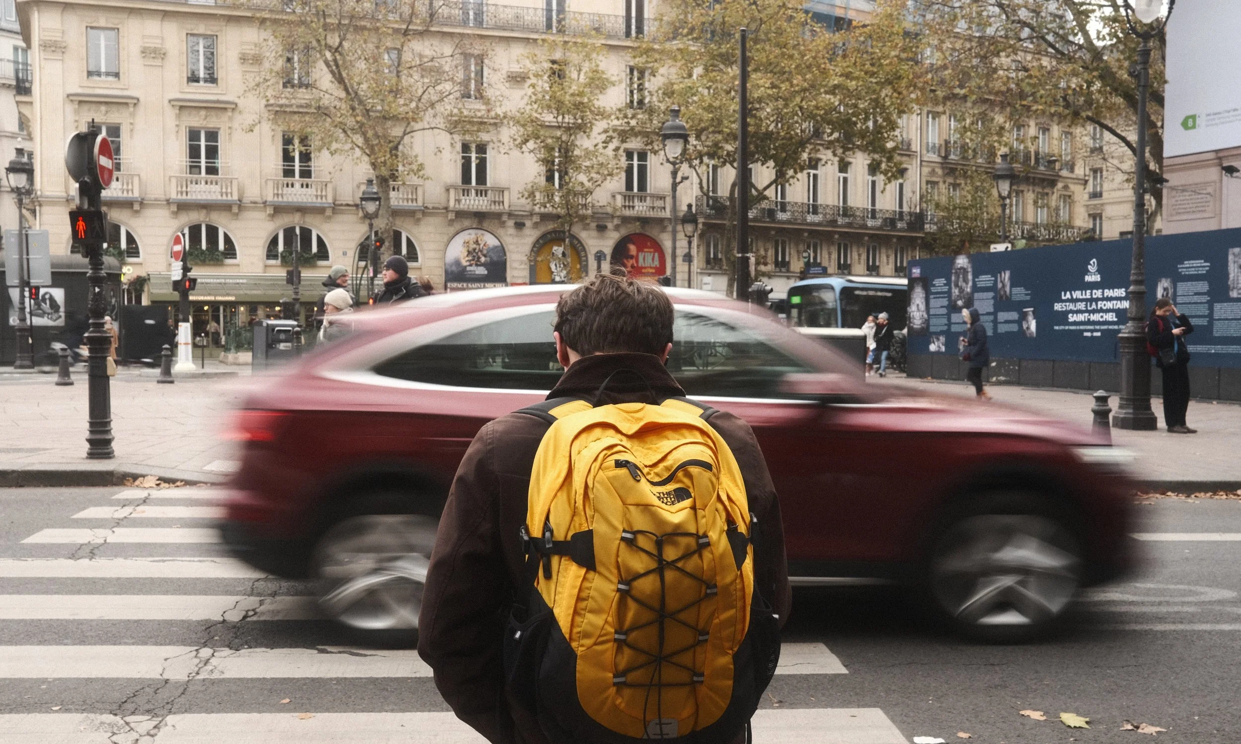 Un homme avec un sac à dos jaune regarde une rue animée à Paris, avec des voitures en mouvement et des passants, en automne.