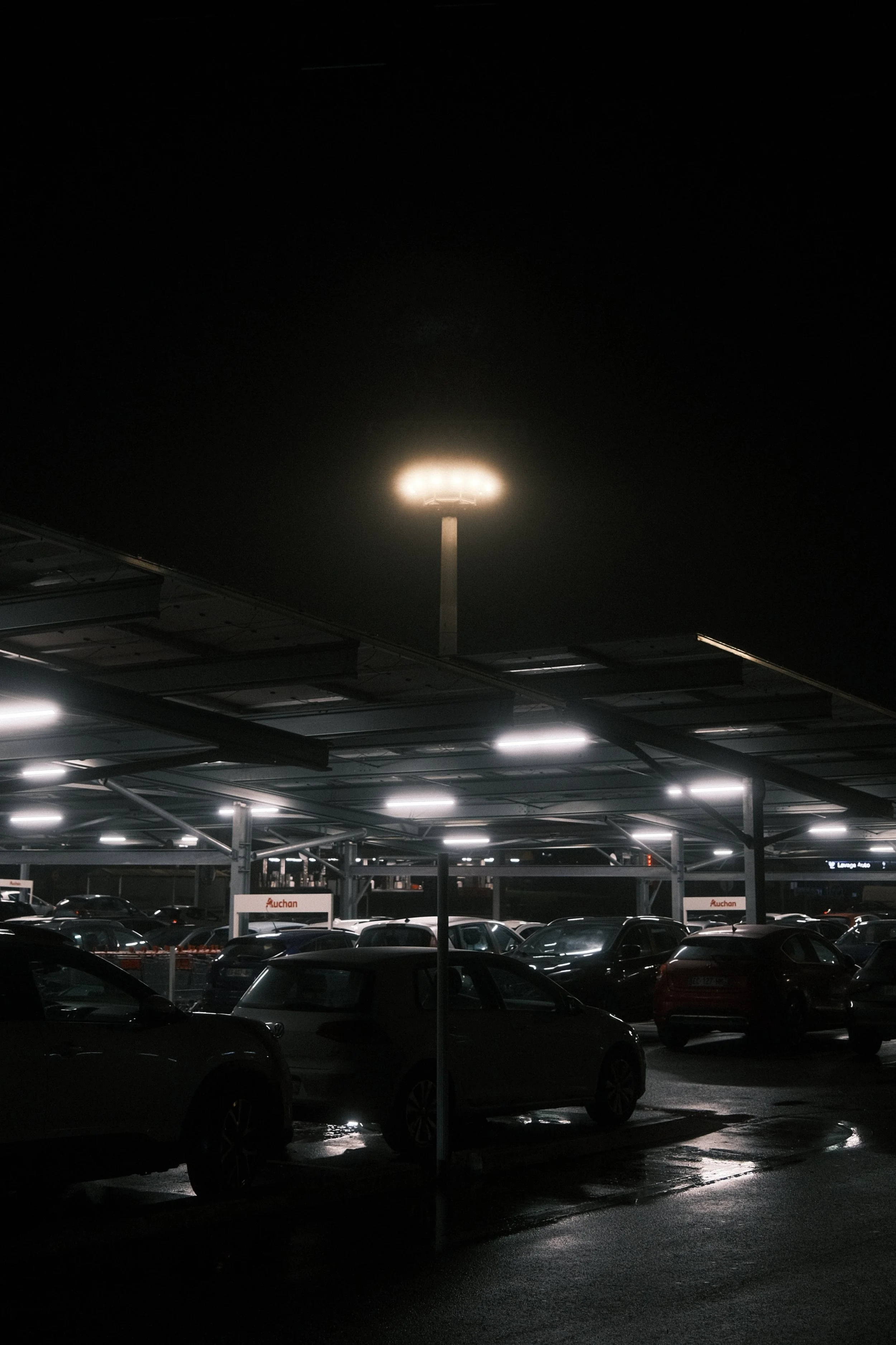 Parking souterrain sous un centre commercial, éclairage artificiel et voitures stationnées, ciel nocturne visible au-dessus.