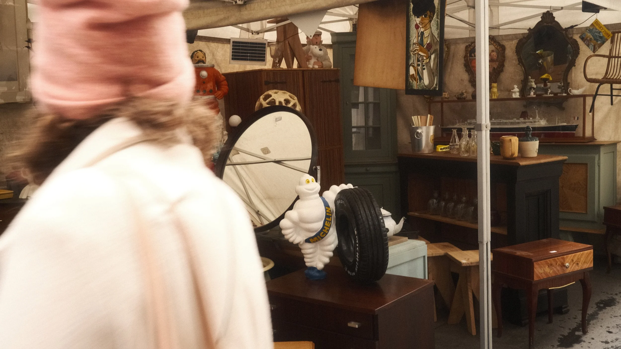 Divers objets de décoration vintage, y compris une figurine de la mascotte Michelin, des meubles anciens et des bibelots. Une personne passe devant avec un flou de mouvement.