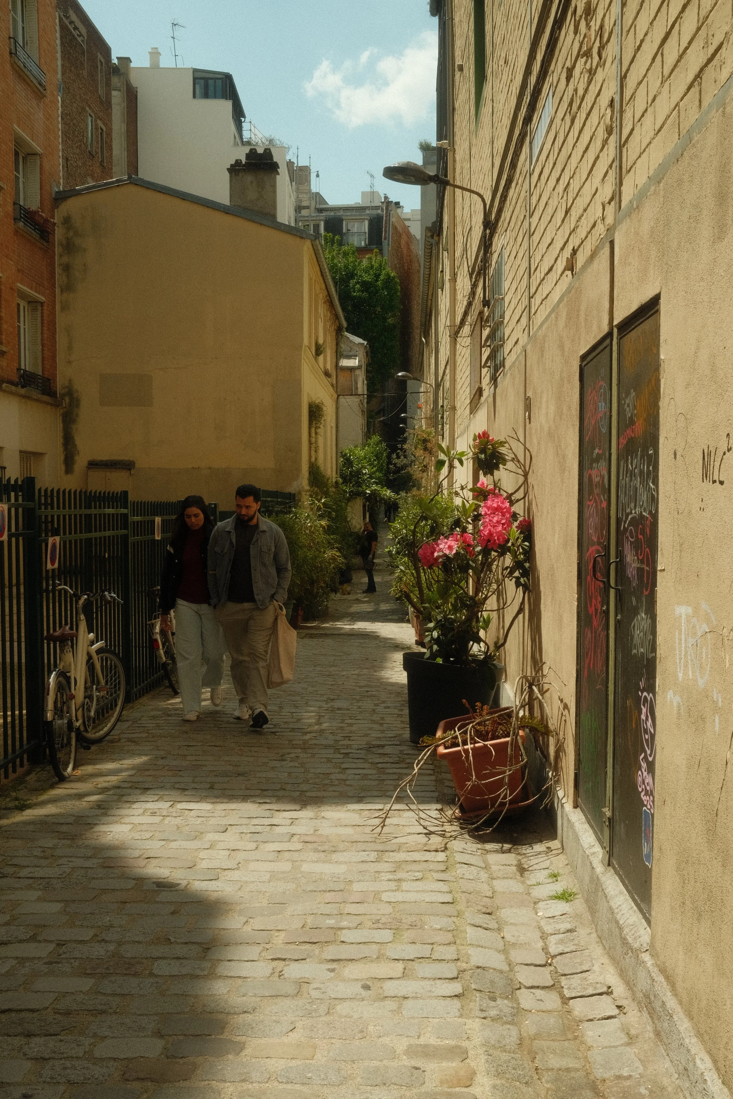 Deux personnes marchent dans une ruelle pavée bordée de bâtiments avec des fleurs en pot sur le côté droit.
