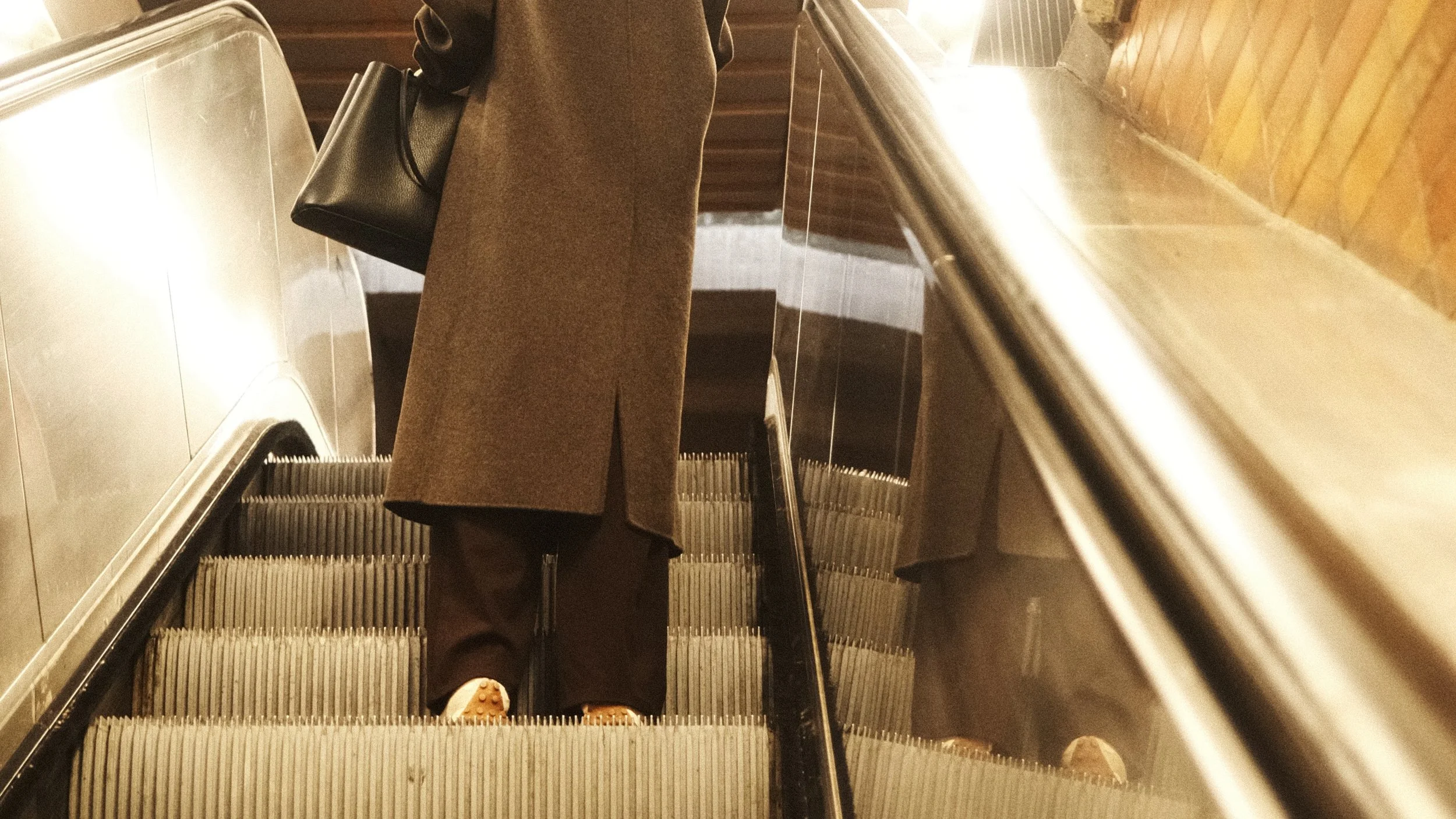 Person montant dans un escalator, portant un manteau marron et un sac noir, avec un mur en briques en arrière-plan.