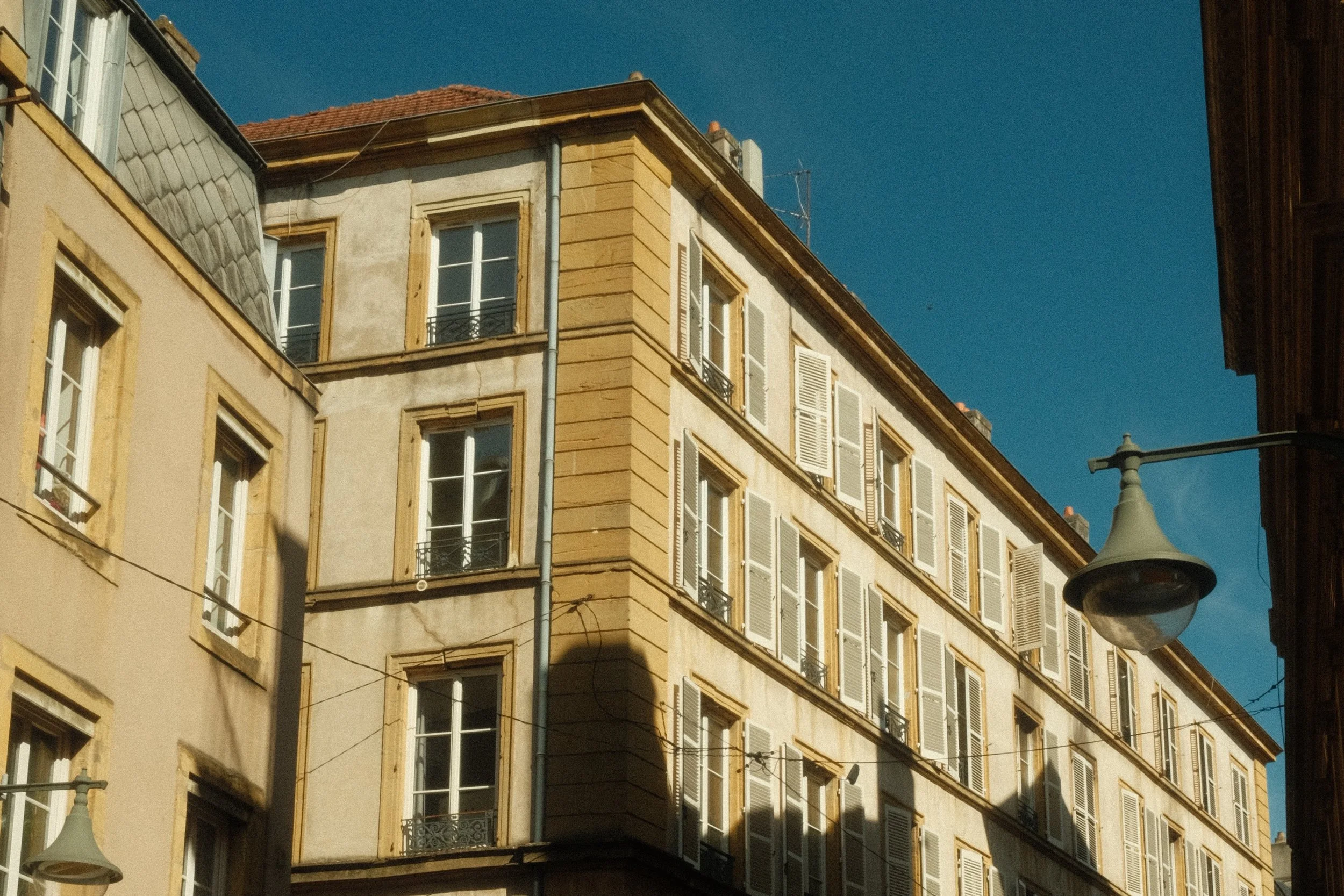 Bâtiment d'architecture classique avec plusieurs fenêtres à volets blancs, face à un ciel bleu, avec un lampadaire en premier plan.