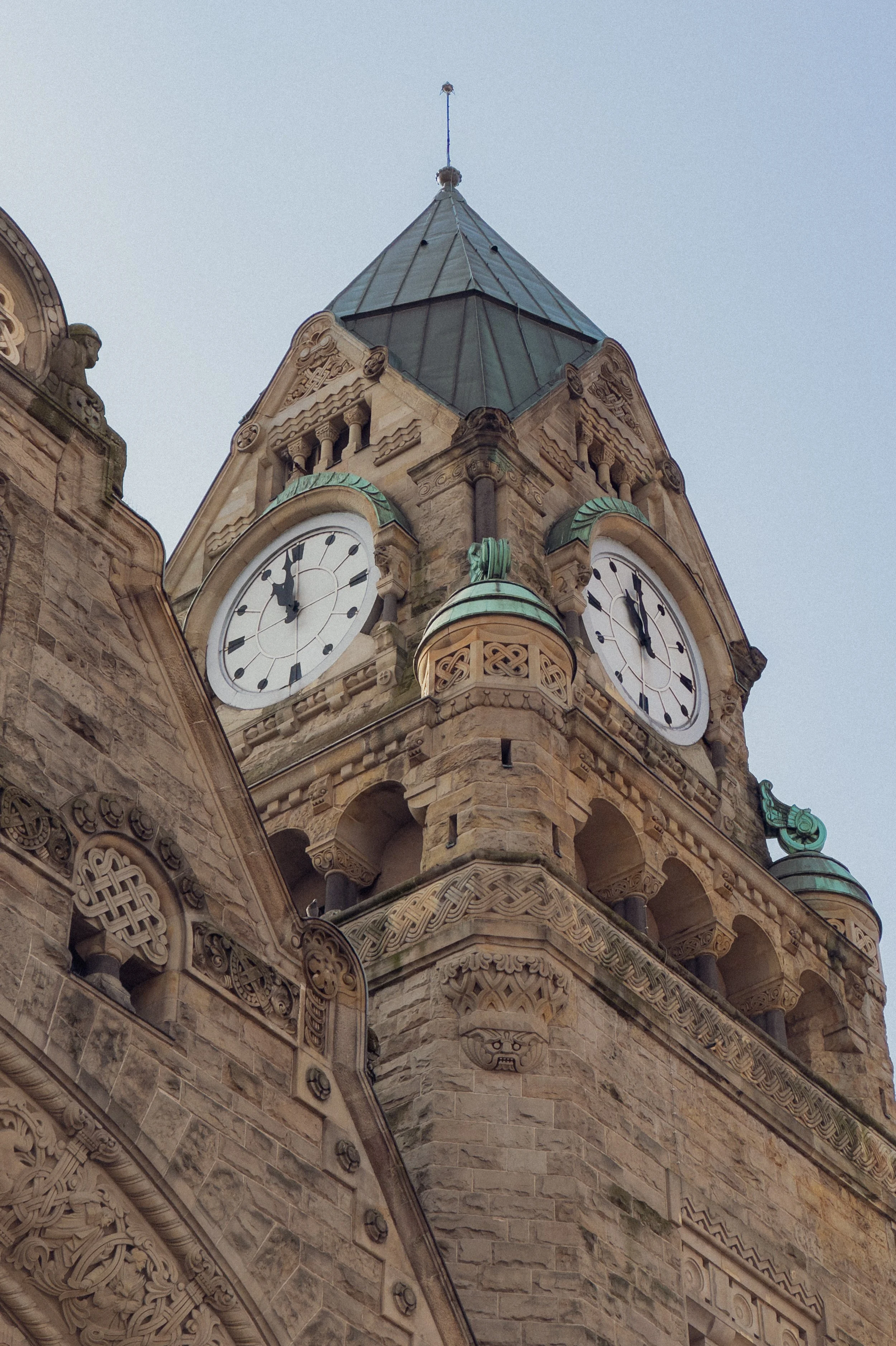 Une tour d'horloge en pierre avec deux cadrans d'horloge, une toiture en métal vert patiné, et des détails architecturaux ornementaux, sous un ciel gris.