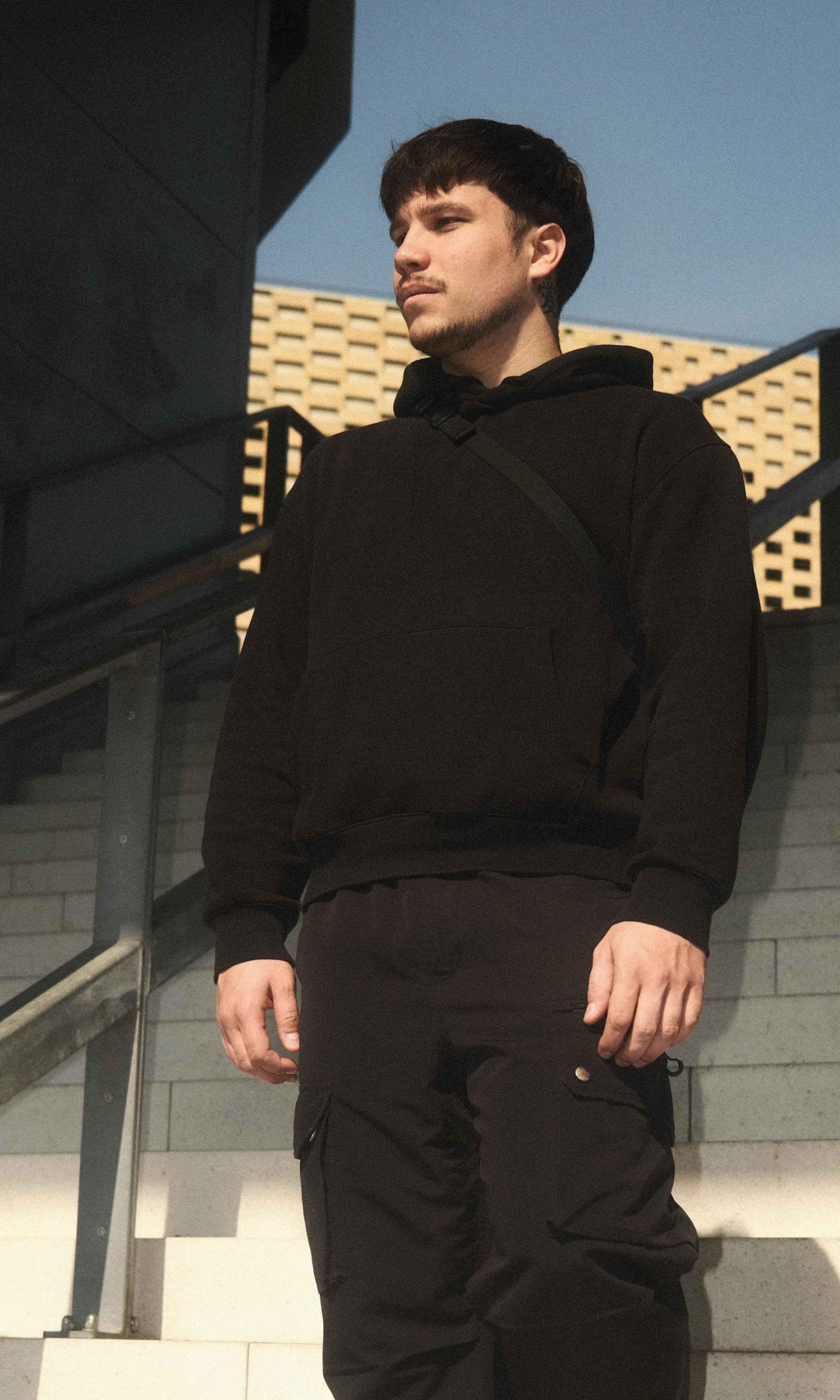 Un jeune homme portant un sweat noir et un pantalon noir, debout près d'une rampe d'escalier en extérieur, avec un bâtiment moderne en arrière-plan et un ciel clair.