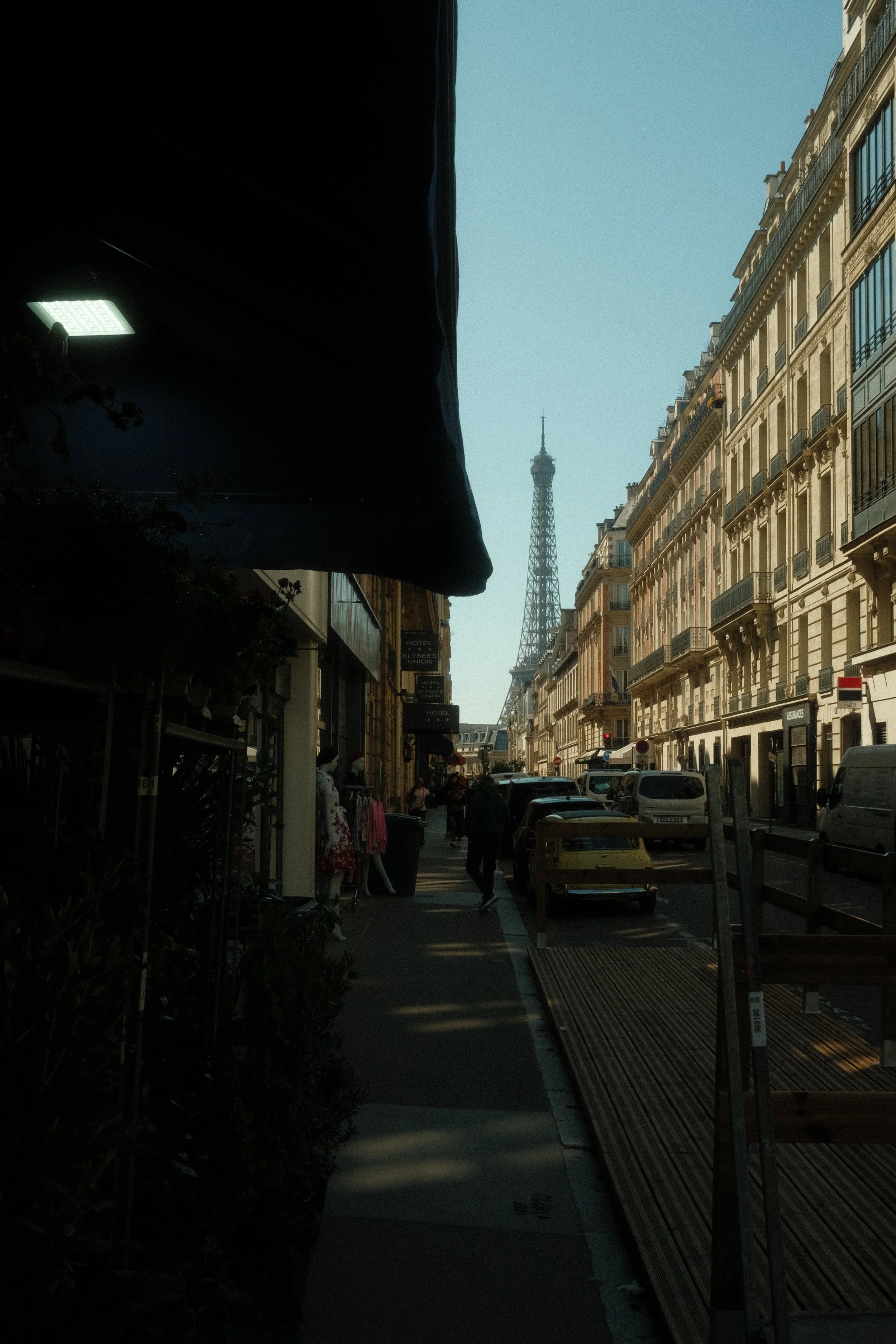 Vue urbaine de Paris avec la tour Eiffel visible au loin, bâtiments haussmanniens et une rue animée sous un ciel bleu clair.