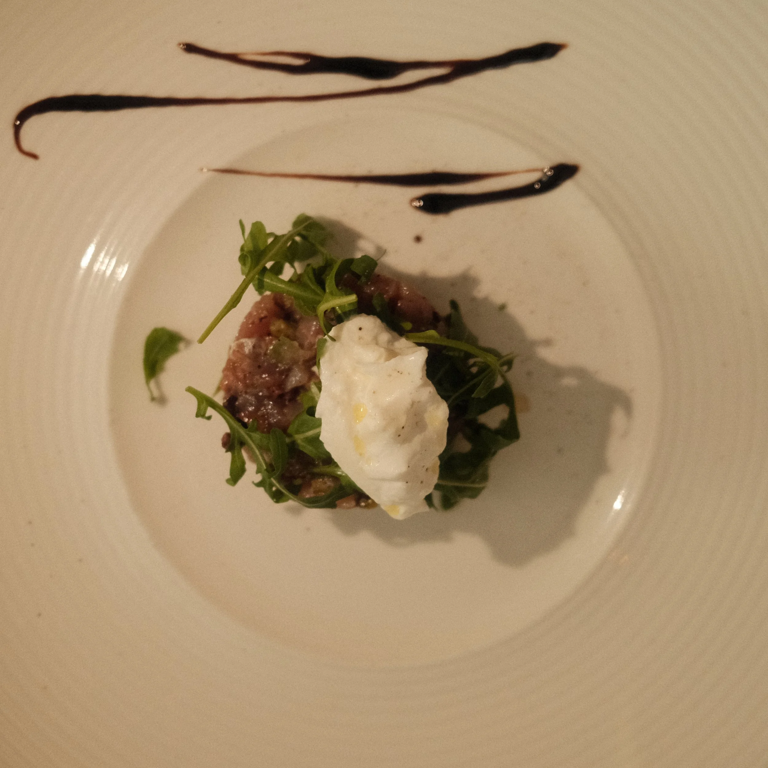 Une assiette avec un morceau de viande, des feuilles vertes, un œuf, de la sauce et des bandes de sauce chocolatées.