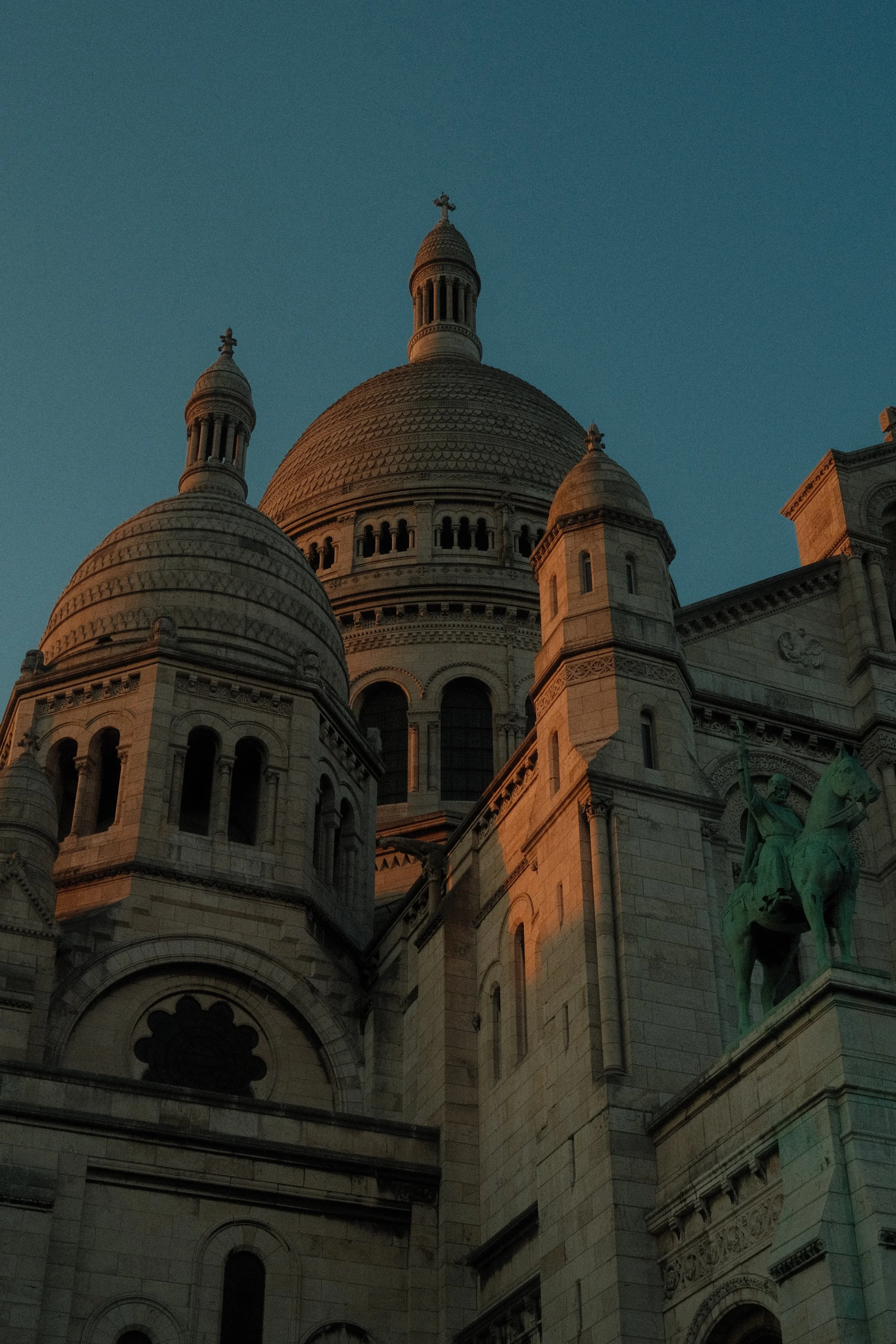 Vue de la basilique du Sacré-Cœur de Paris au coucher du soleil, avec ses dômes et une statue équestre visible à droite.