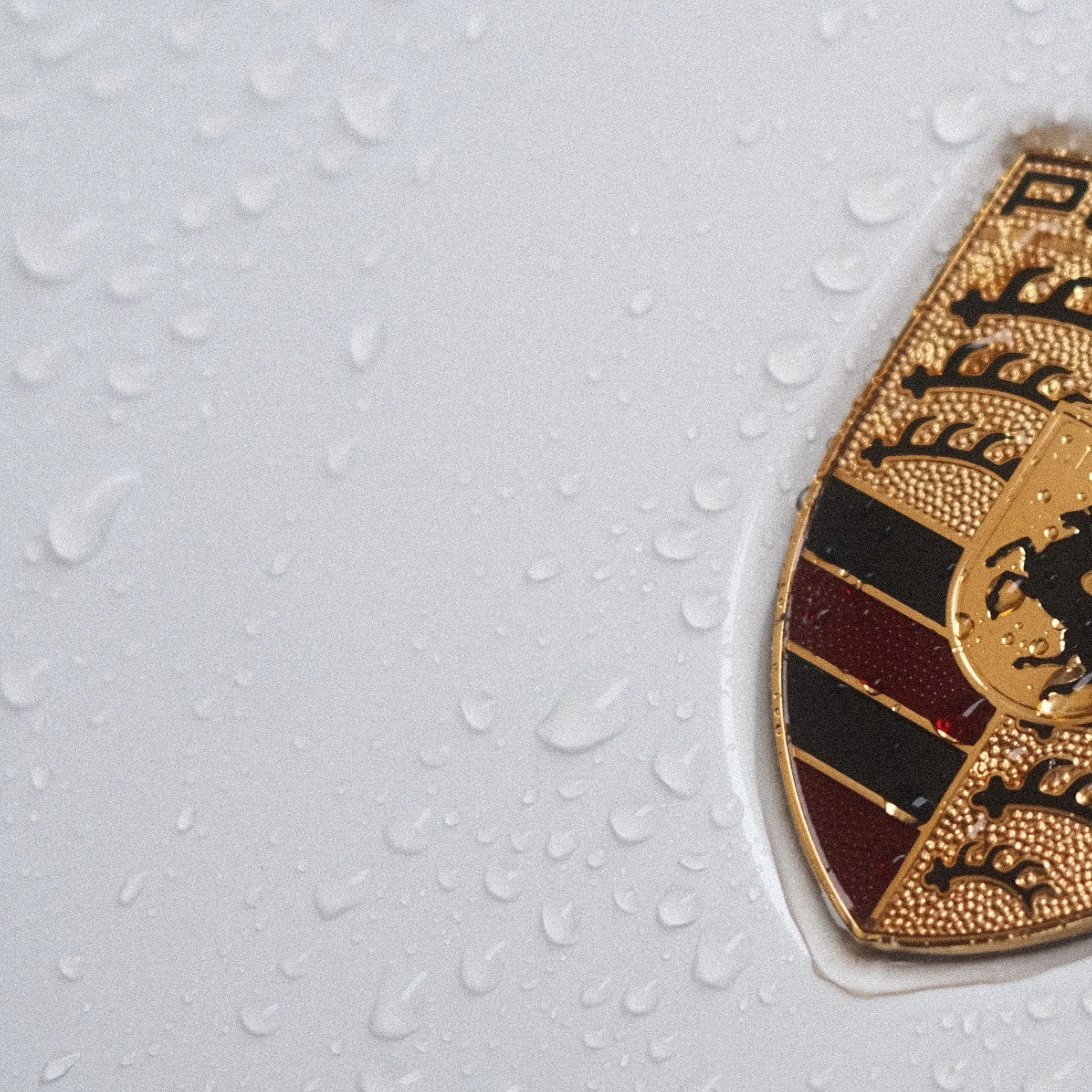 Insigne de Porsche, orné de détails en or noir et rouge, posé sur une surface en plastique avec des gouttes d'eau.