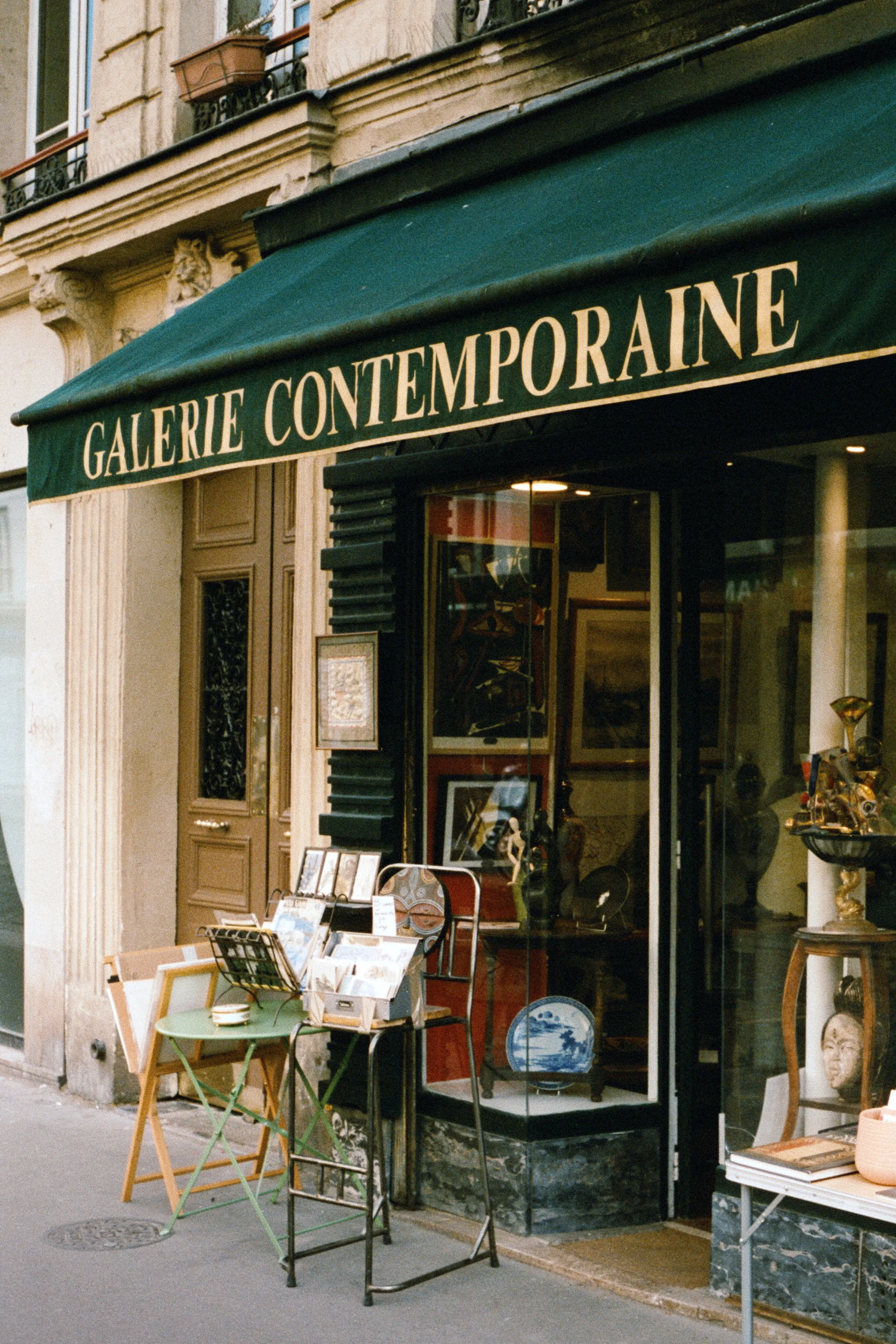 Une galerie d'art appelée 'Galerie Contemporaine' avec des tableaux exposés à l'intérieur et des œuvres d'art visibles à travers la vitrine, avec une étagère extérieure présentant des livres ou des cartes.
