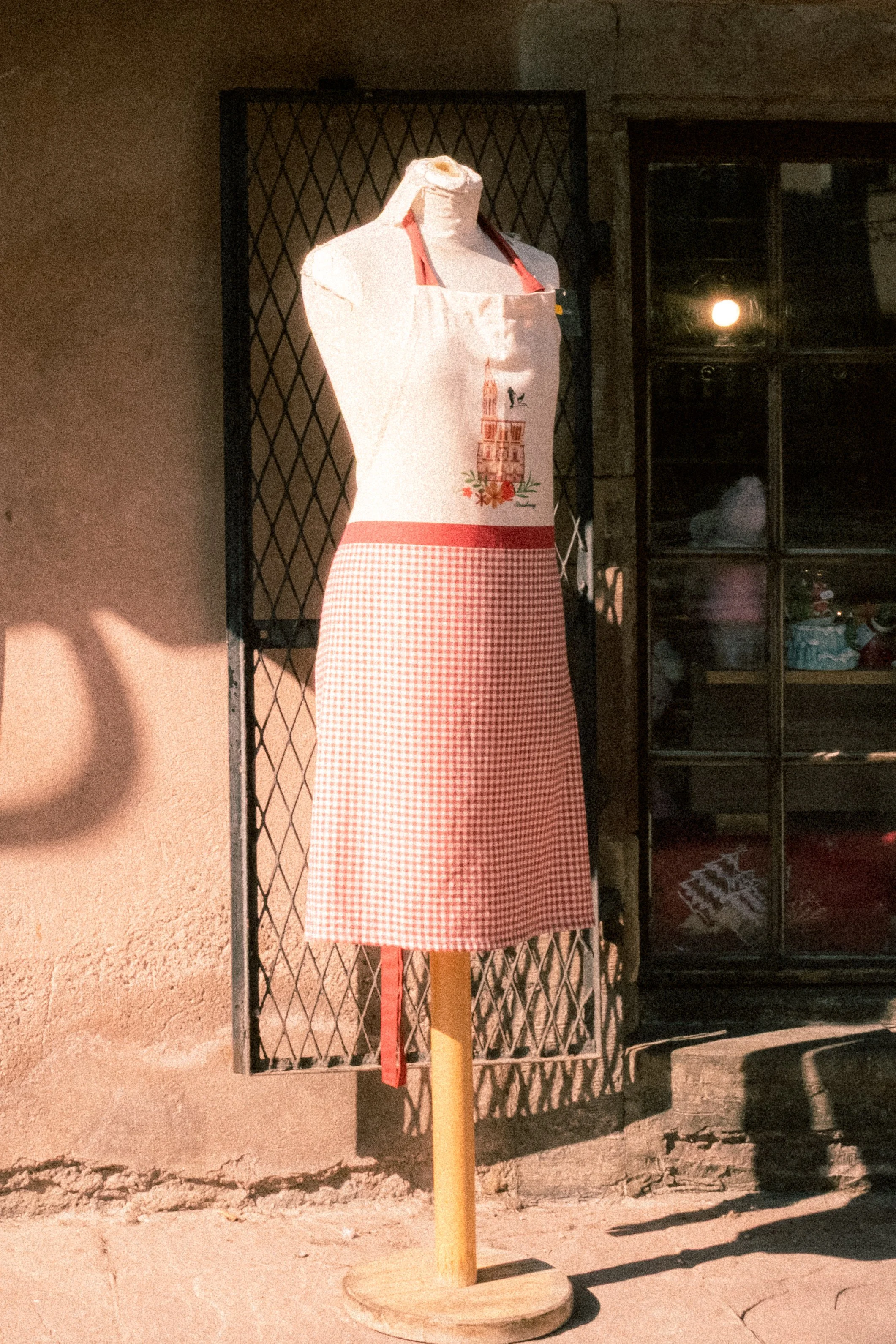 Mannequin portant un tablier rouge et blanc à carreaux, affiché en extérieur près d'un mur et d'une fenêtre.