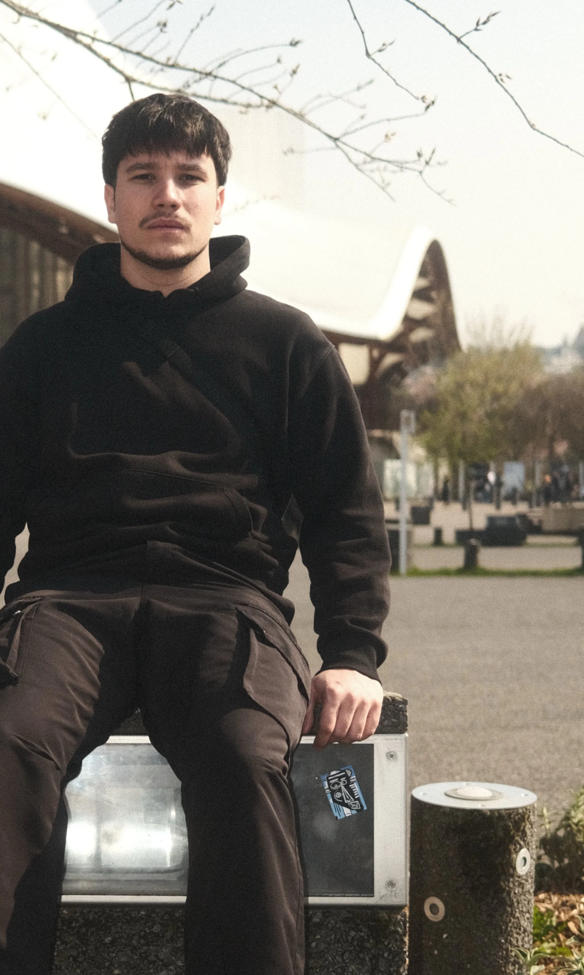 Jeune homme assis sur un banc en plein air, portant un sweat noir et un pantalon beige, avec un bâtiment moderne en arrière-plan et des branches d'arbre nuves dans le ciel.