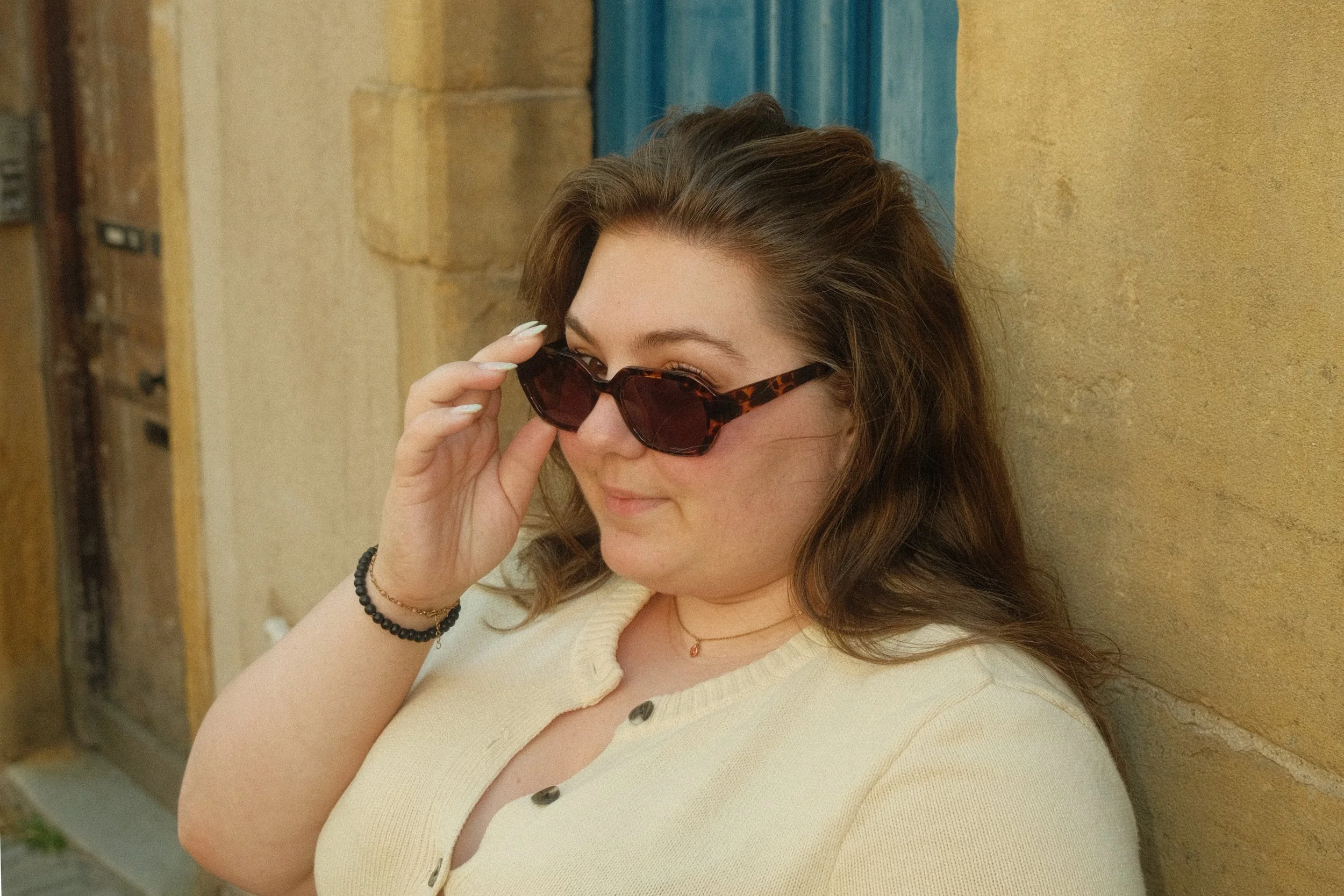 Jeune femme portant des lunettes de soleil à motif tortue, tenant la main à la monture, en avant-plan, avec un mur beige en arrière-plan.