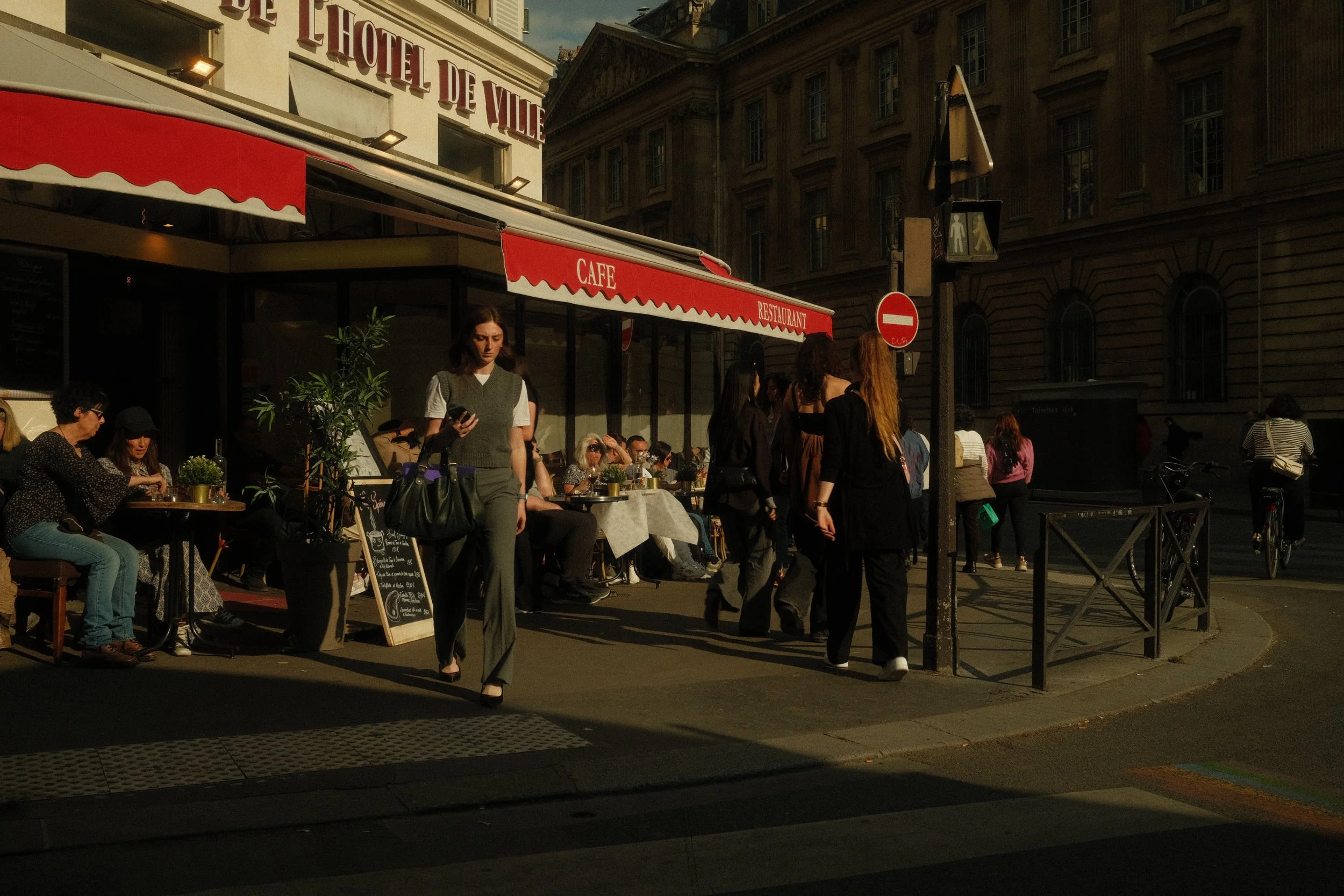 Une scène urbaine devant un café avec des gens assis et debout, une enseigne de café, et un bâtiment historique en arrière-plan, le tout en lumière de fin d'après-midi.