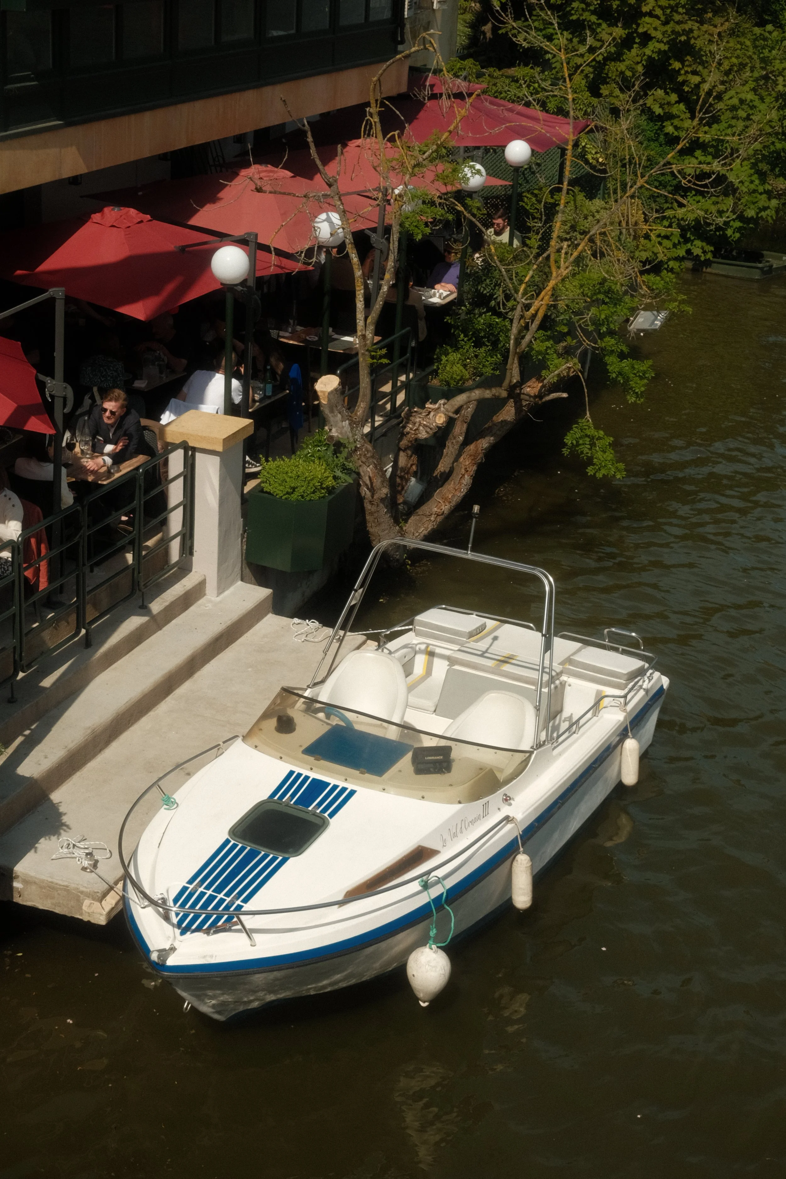 Un bateau blanc amarré au quai près d'une terrasse de restaurant en bord d'eau, avec des parasols rouges et des clients assis à des tables. Il y a un arbre avec des branches qui s'étendent au-dessus du quai.