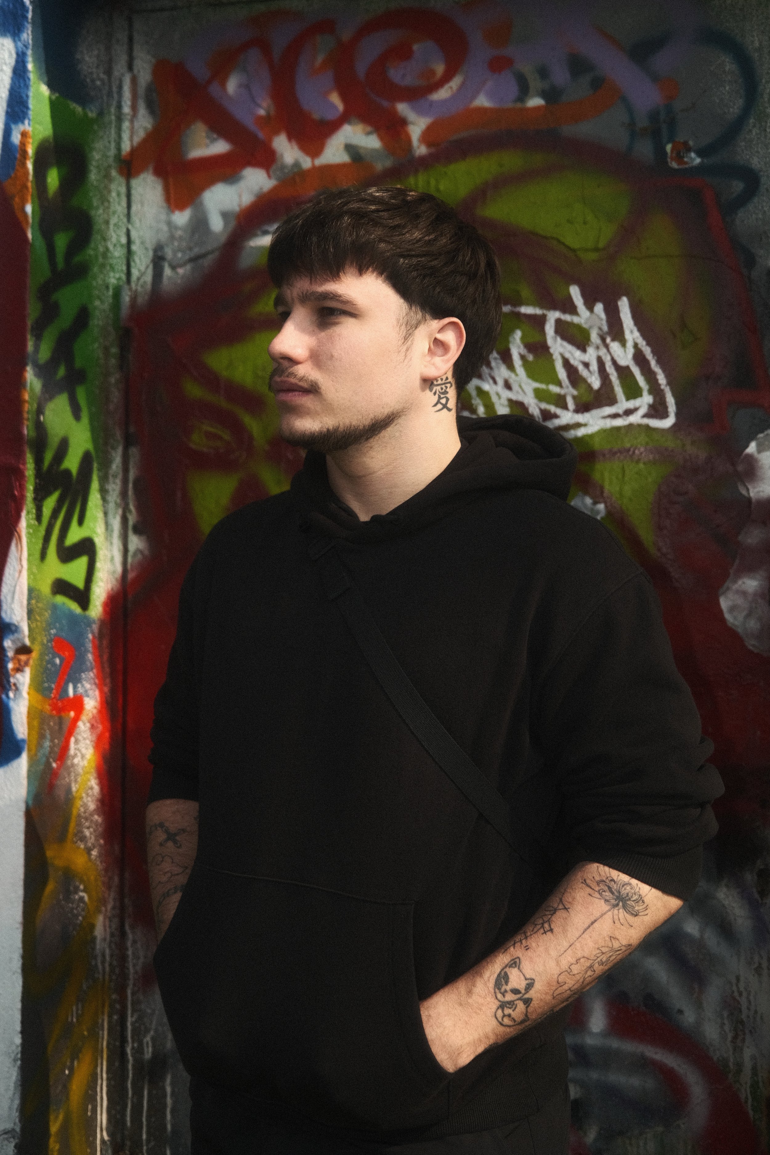 Jeune homme avec un tatouage au cou, portant un sweat noir, debout devant un mur de graffitis colorés.