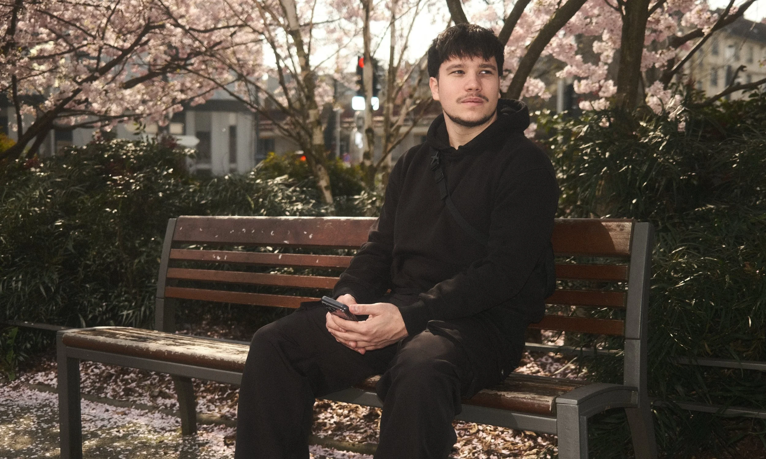 Jeune homme assis sur un banc dans un parc, portant un hoodie noir, tenant un téléphone dans ses mains, avec des fleurs de cerisier en fleurs en arrière-plan.