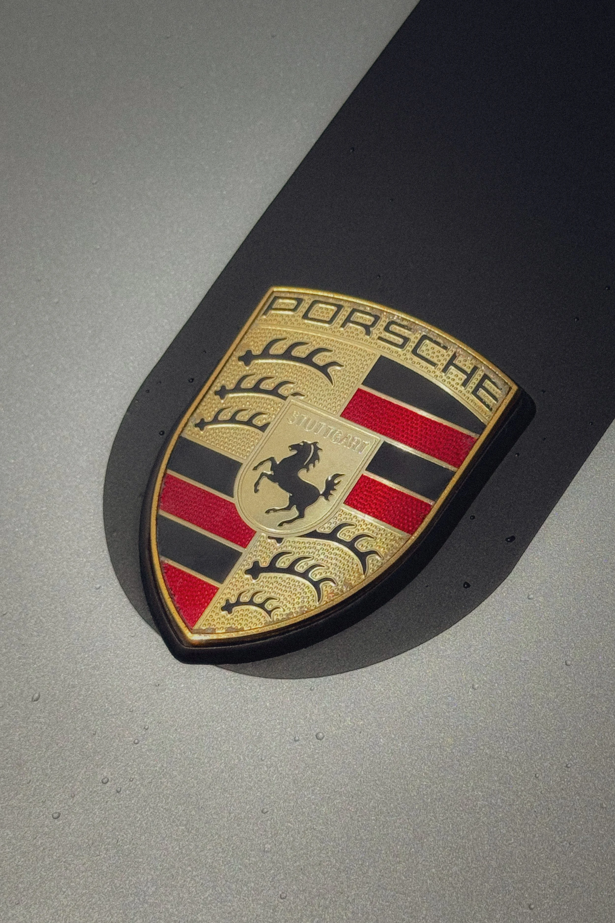 Logo de la marque Porsche sur le capot d'une voiture, avec une seule ombre portée.