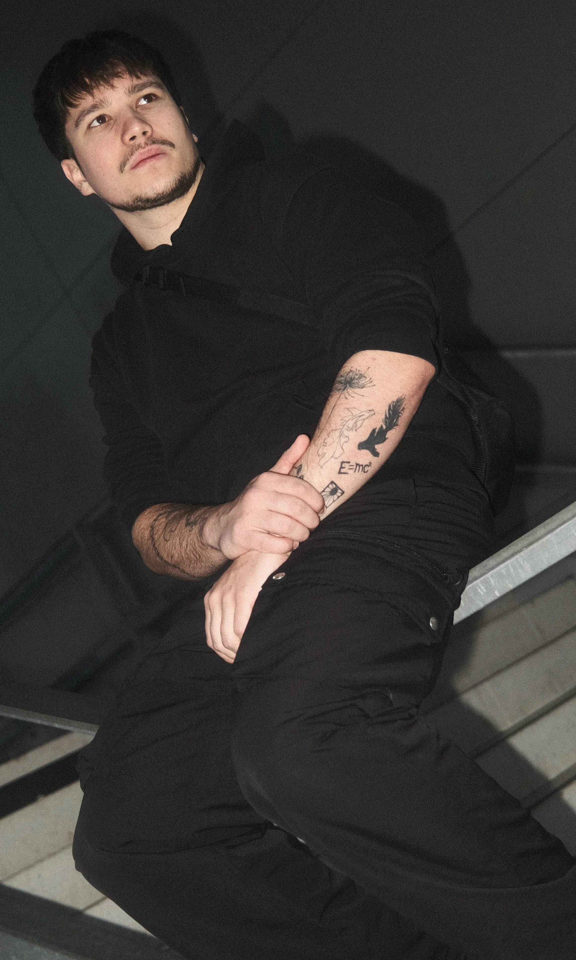 Jeune homme assis, portant un sweat noir, avec des tatouages visibles sur le bras, dans un environnement sombre.