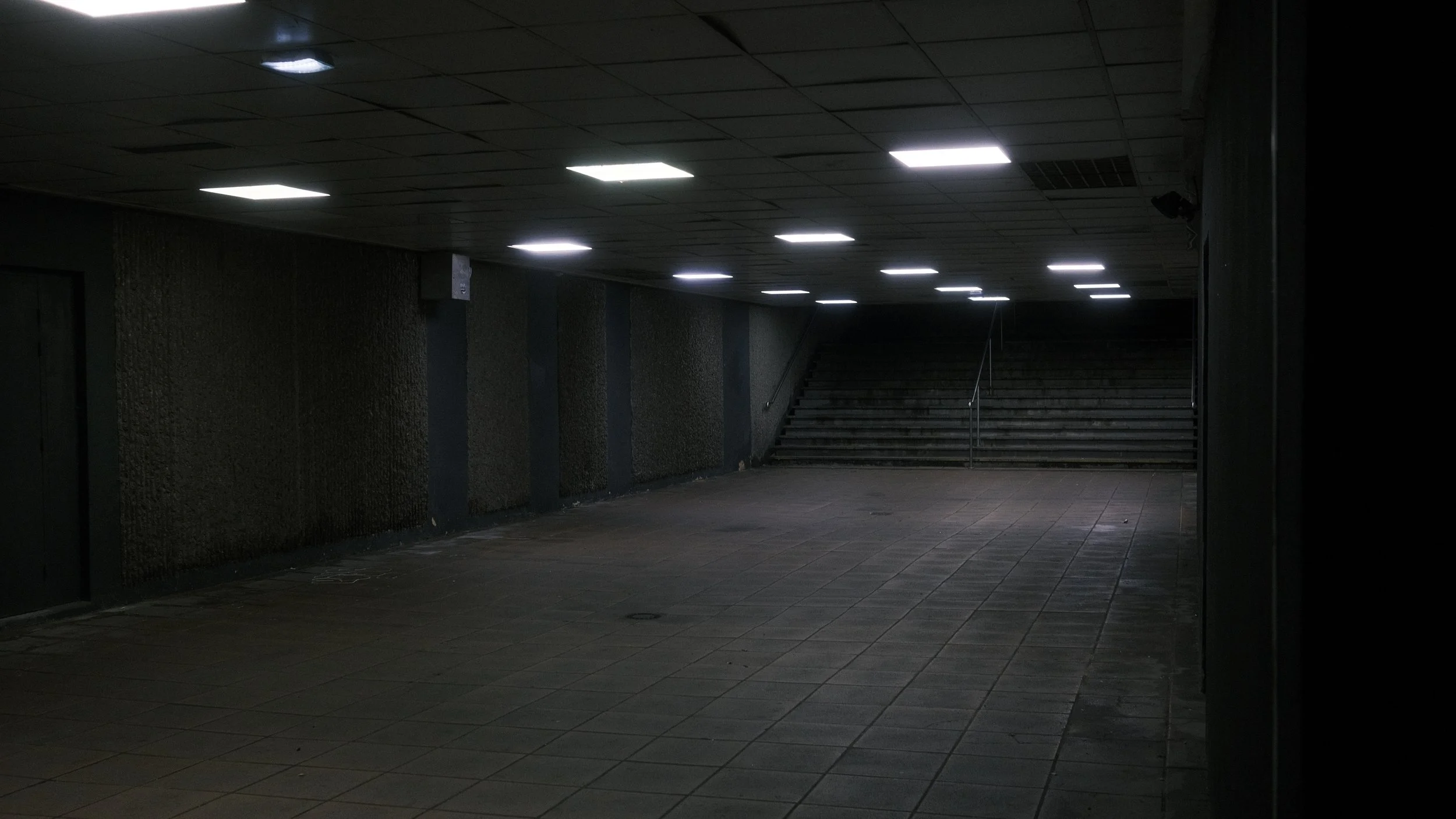 Passage souterrain sombre avec éclairage fluorescent, escaliers à l'extrémité et murs en béton.