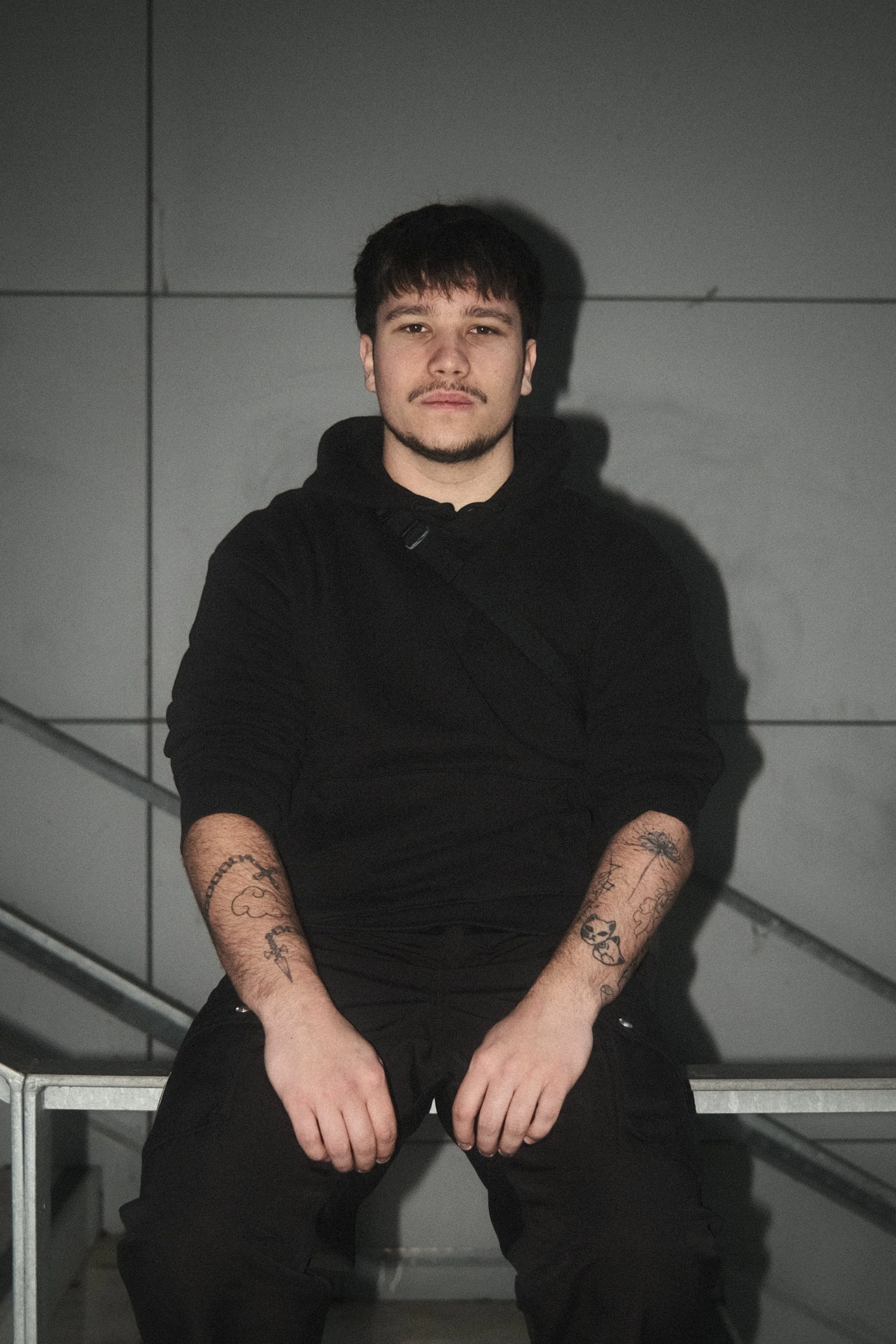 Un jeune homme assis sur un banc, portant un pull noir et des tatouages visibles sur les avant-bras, dans un environnement urbain avec un mur gris en arrière-plan.