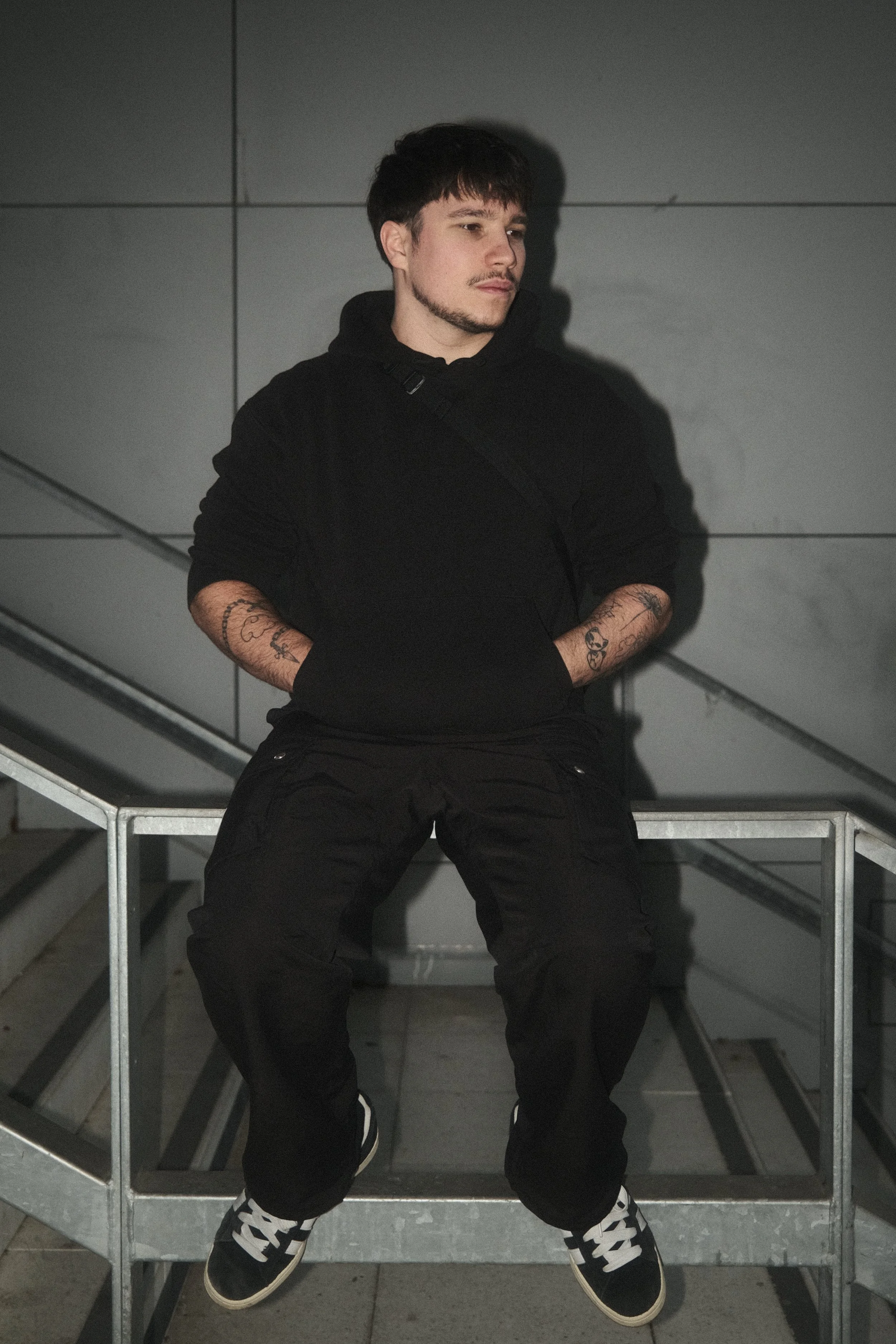 Jeune homme assis sur une surface en métal, portant un sweat à capuche noir, pantalon cargo noir, et des chaussures noires et blanches, dans un environnement urbain.