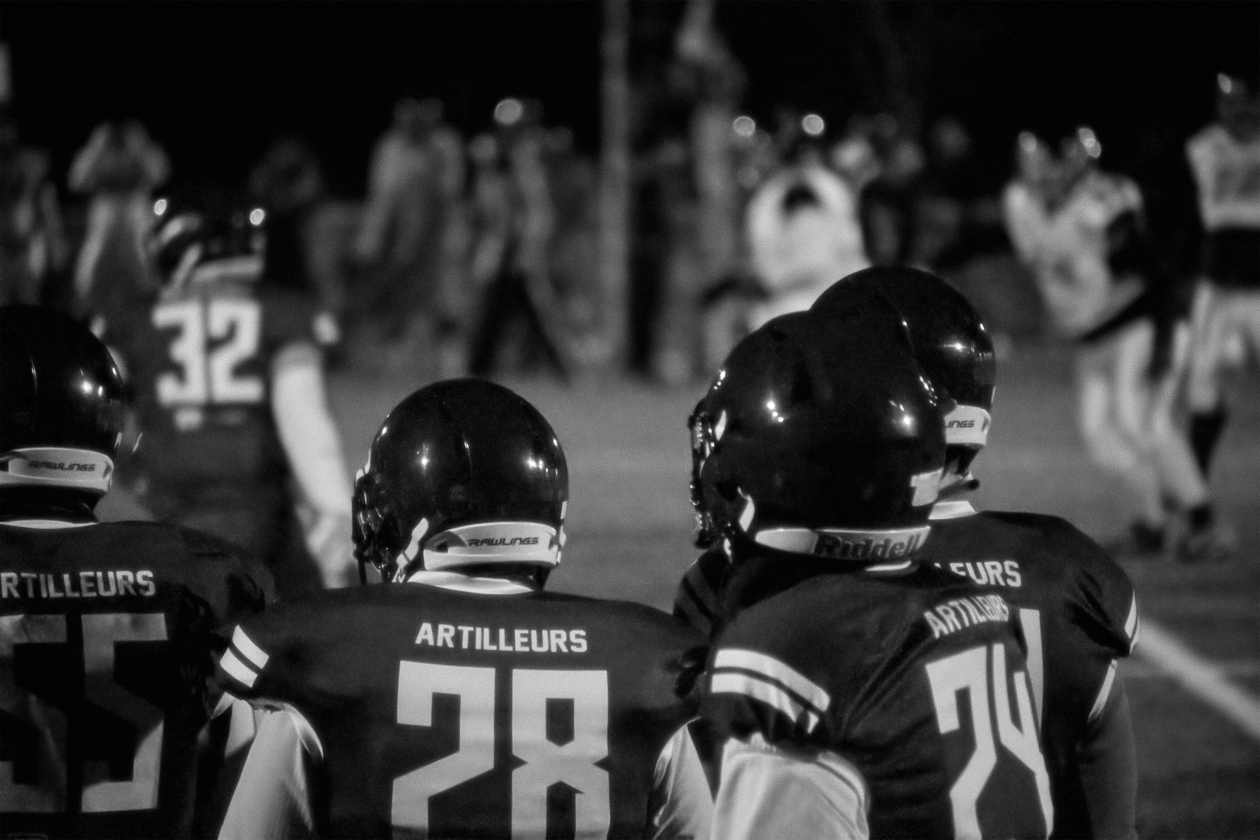 Photo de nuit montrant des joueurs de football américain en uniforme, portant des casques noirs, avec le mot 'ARTILLEURS' et des numéros visibles sur leurs maillots. Des joueurs sont en position défensive, préparant pour la prochaine action, avec des