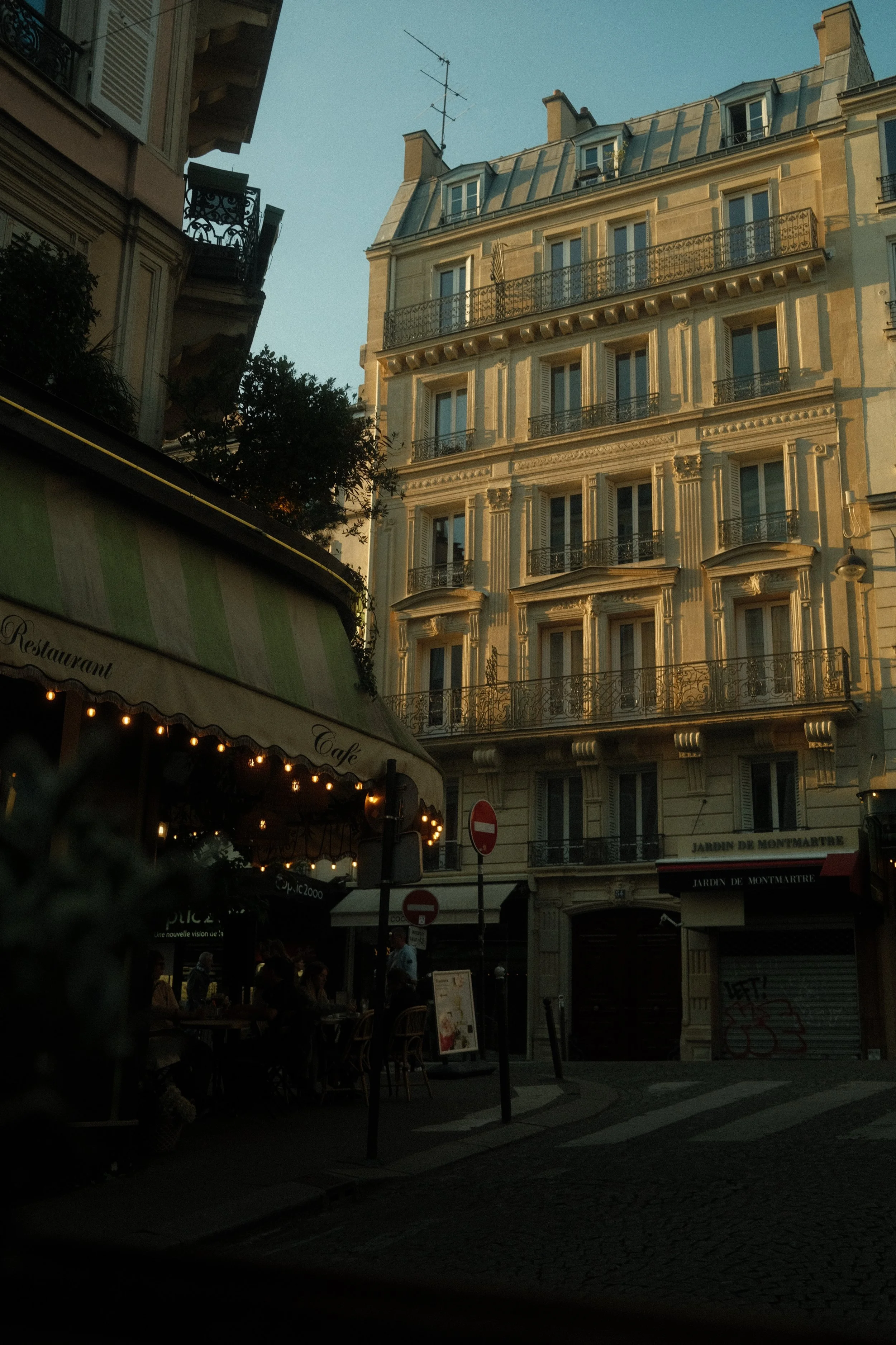 Une rue parisienne avec un bâtiment haussmannien à plusieurs étages, un restaurant en plein air avec des lumières décoratives et un ciel bleu au crépuscule.