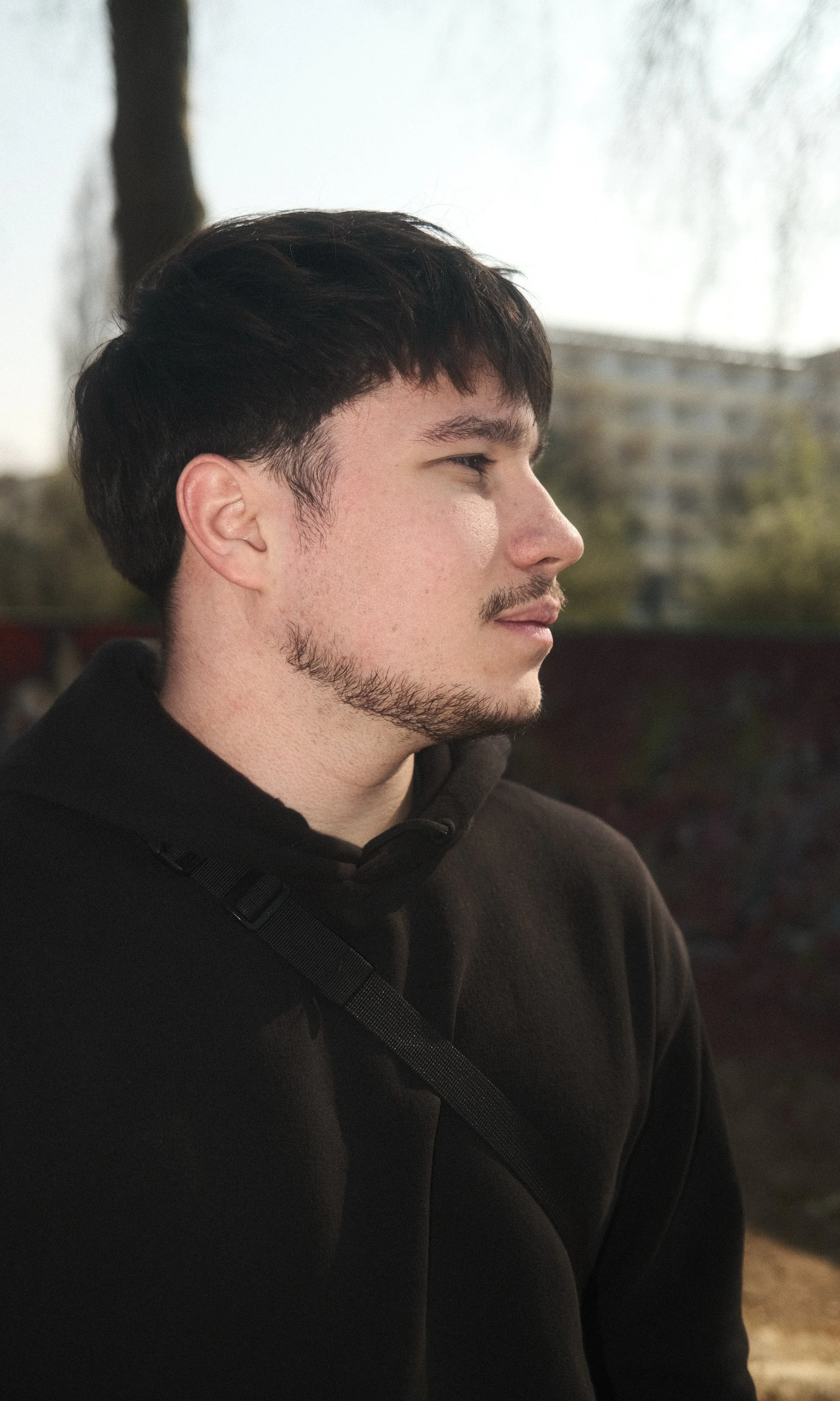 Portrait d'un jeune homme de profil avec une barbe, portant un sweat noir, en extérieur, avec un arbre et un bâtiment en arrière-plan.