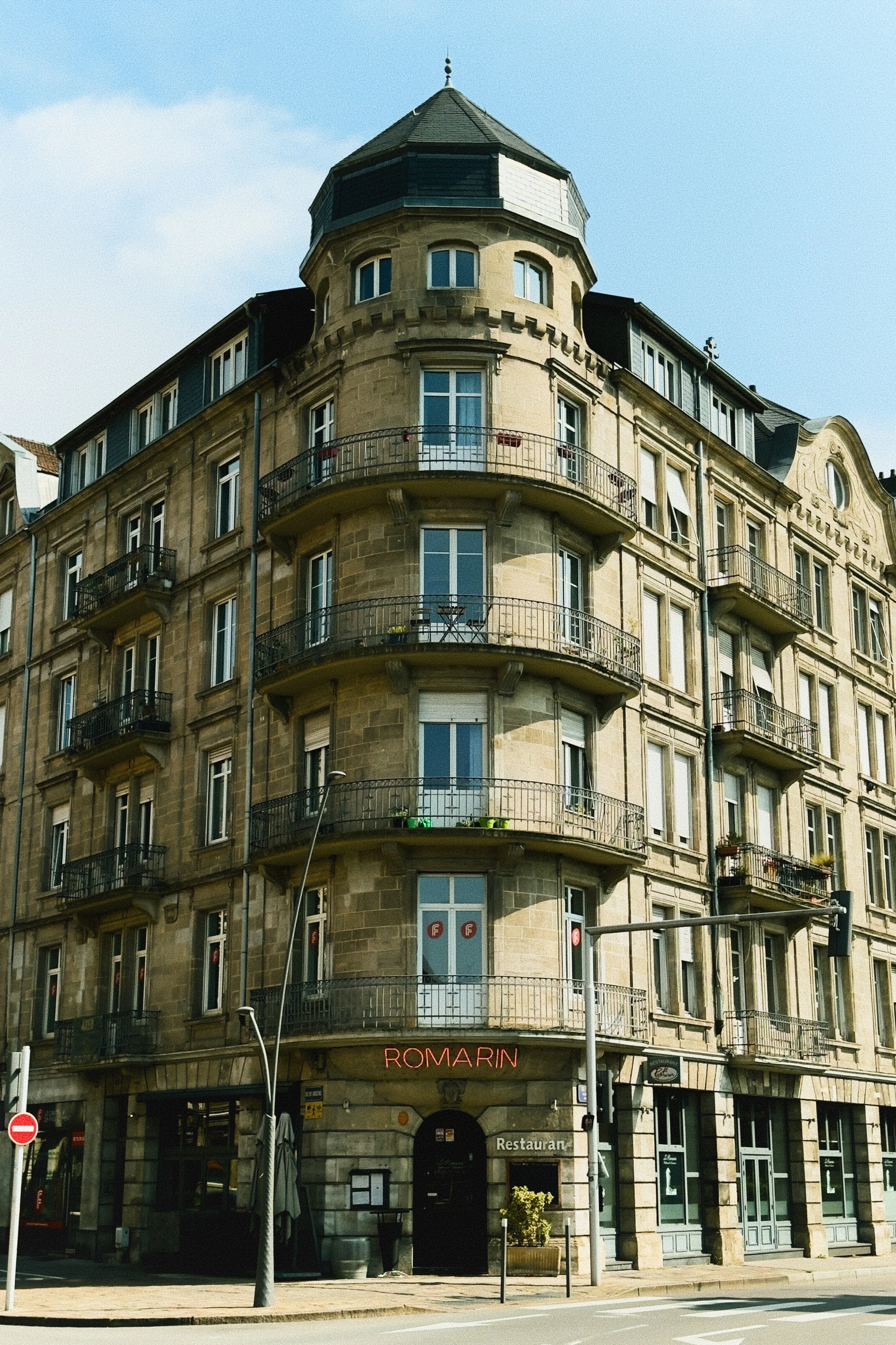 Bâtiment ancien à plusieurs étages avec tour en coin, balcons en fer forgé et restaurant "Romarin" au rez-de-chaussée, sous un ciel bleu.