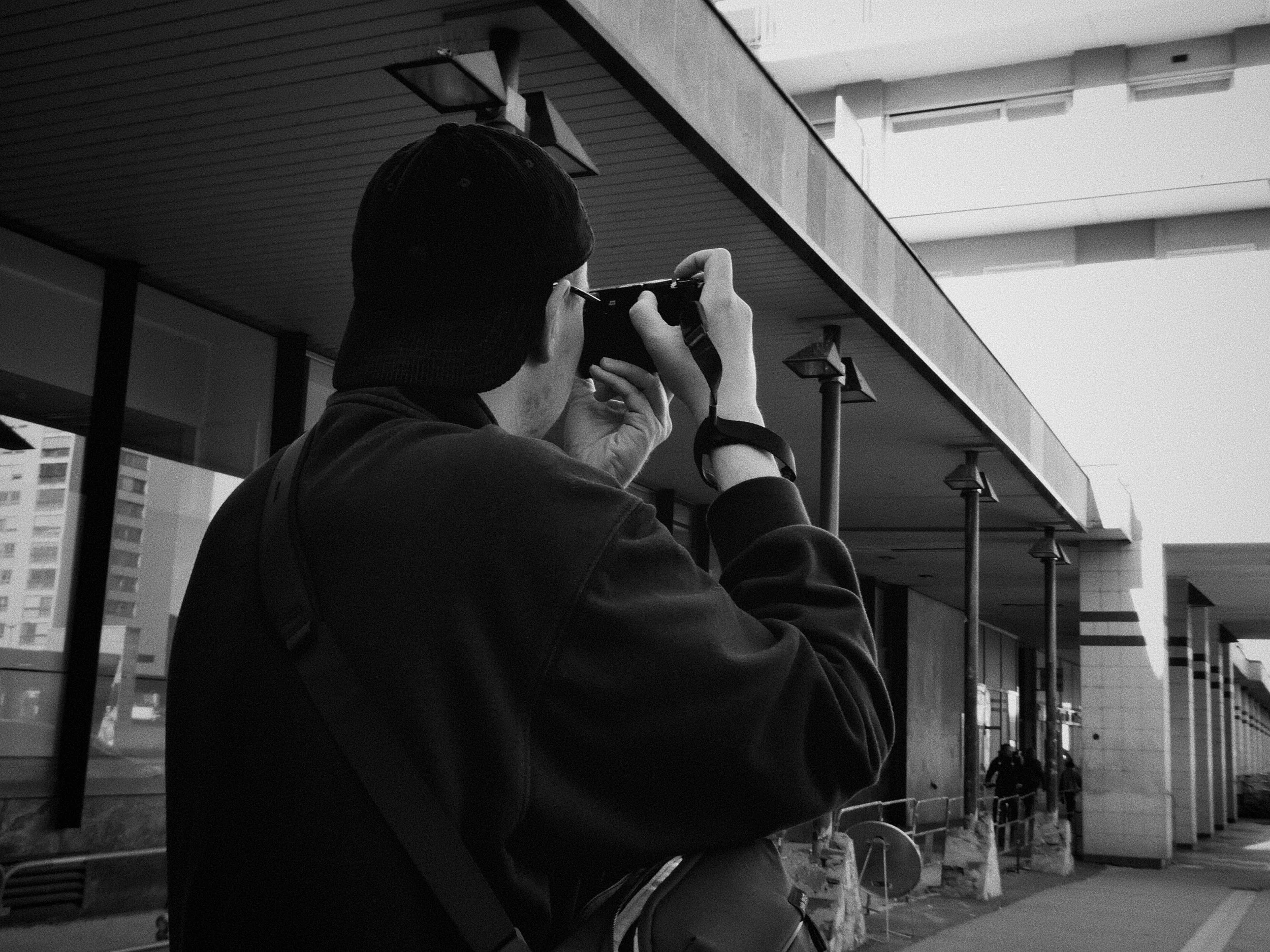 Jeune homme portant un bonnet noir, prenant une photo avec un appareil photo dans un environnement urbain.