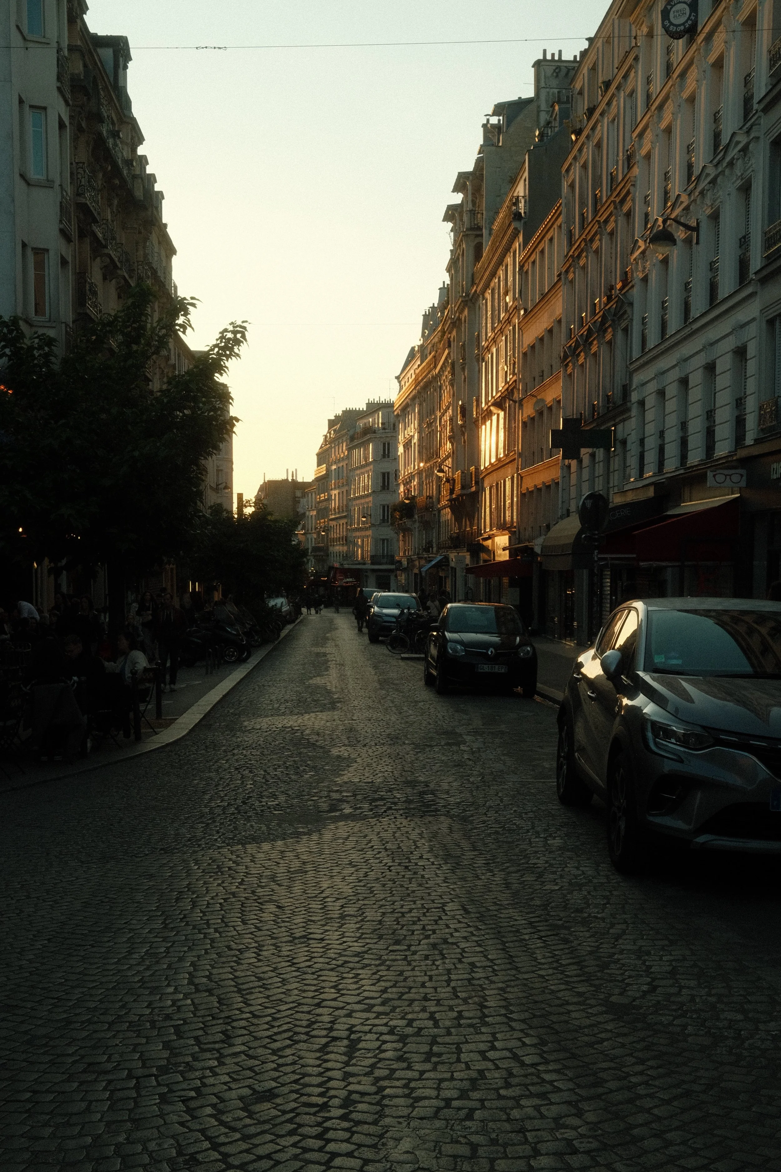 Une rue pavée bordée de bâtiments haussmanniens avec des voitures stationnées et des gens assis à l'extérieur de cafés, sous un ciel au coucher du soleil.