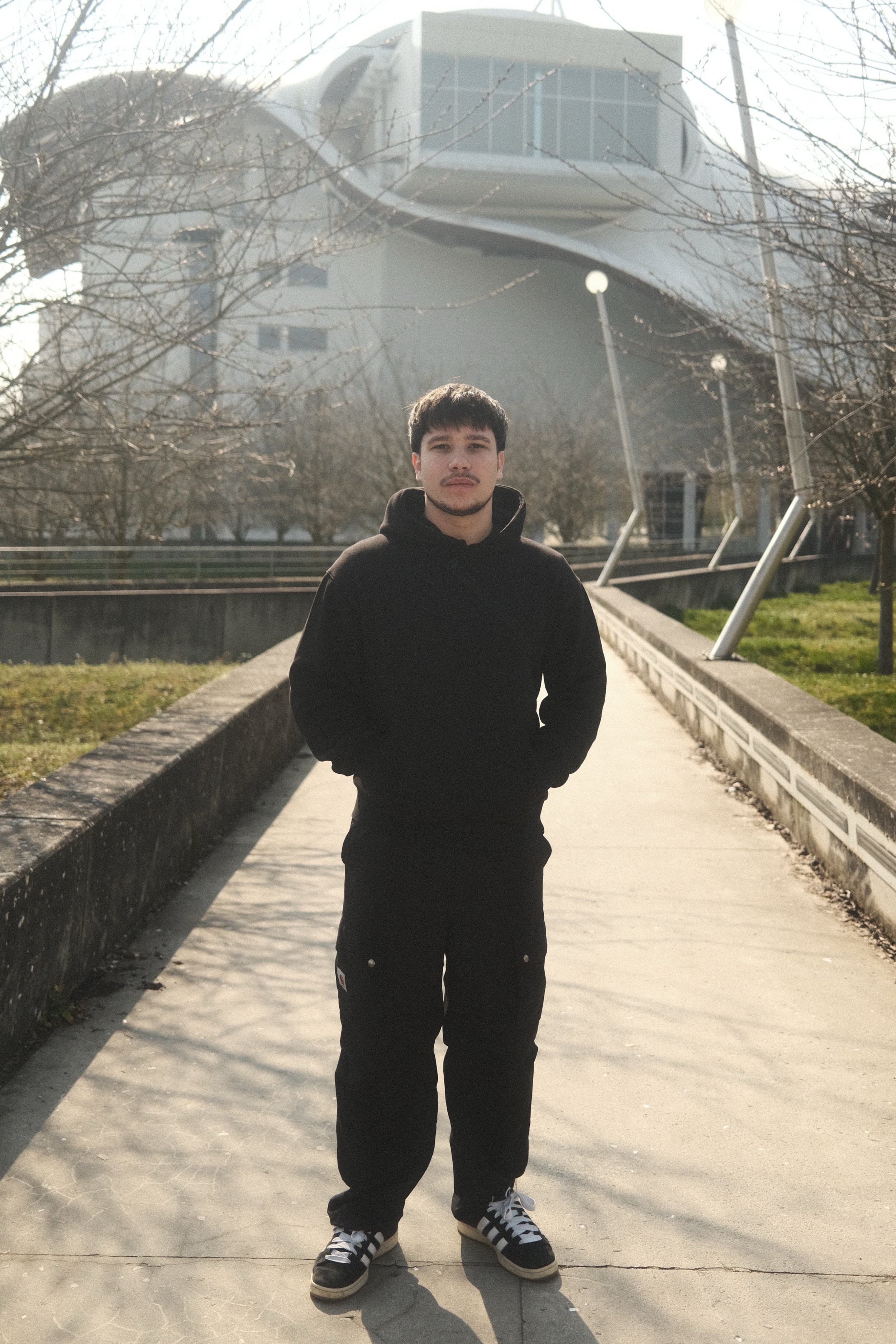 Jeune homme en vêtements décontractés, debout sur un chemin, avec un bâtiment futuriste en arrière-plan, par une journée ensoleillée.