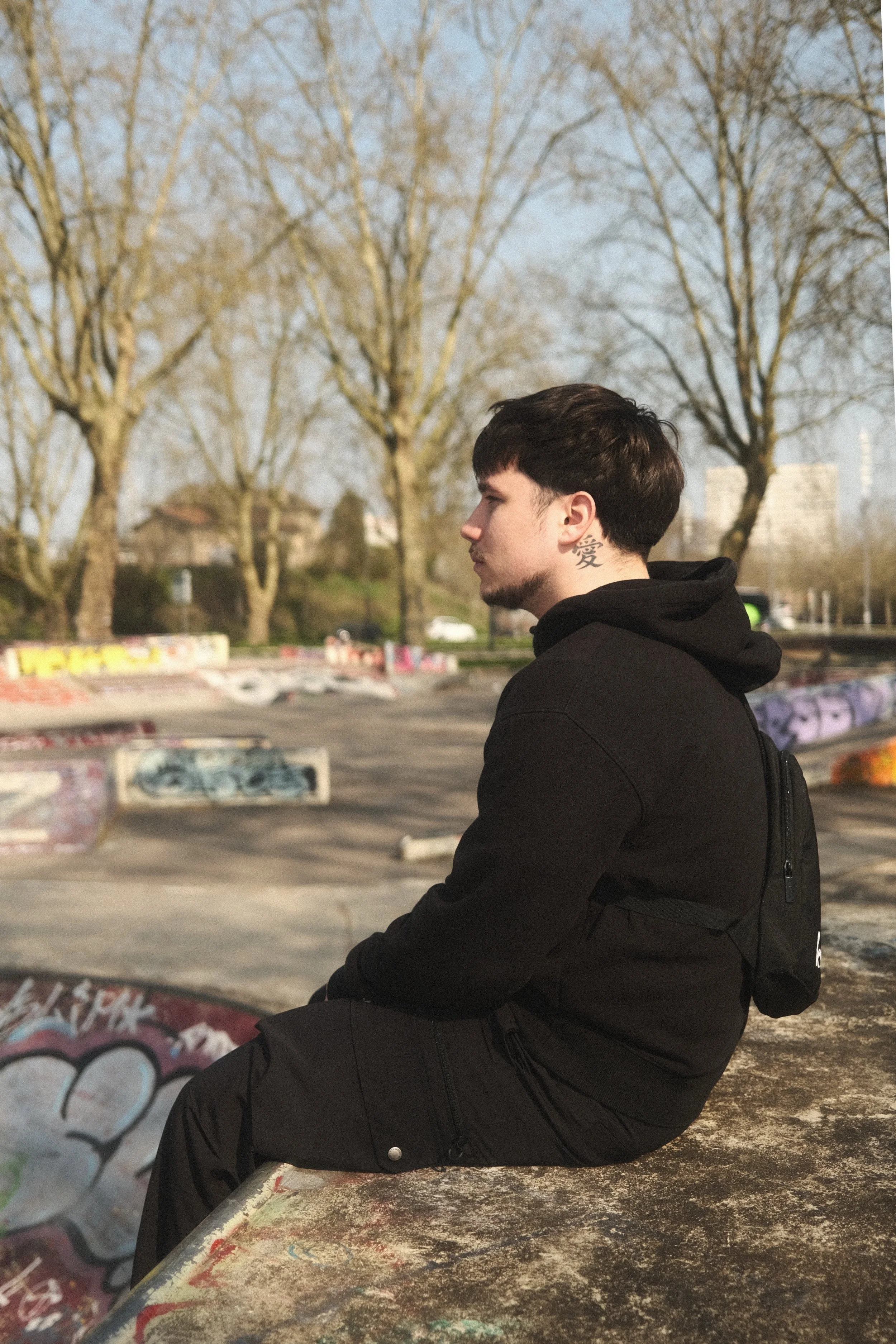 Jeune homme assis sur le bord d'un skatepark avec arbres et graffitis en arrière-plan, portant un sweat noir et un sac à dos noir.