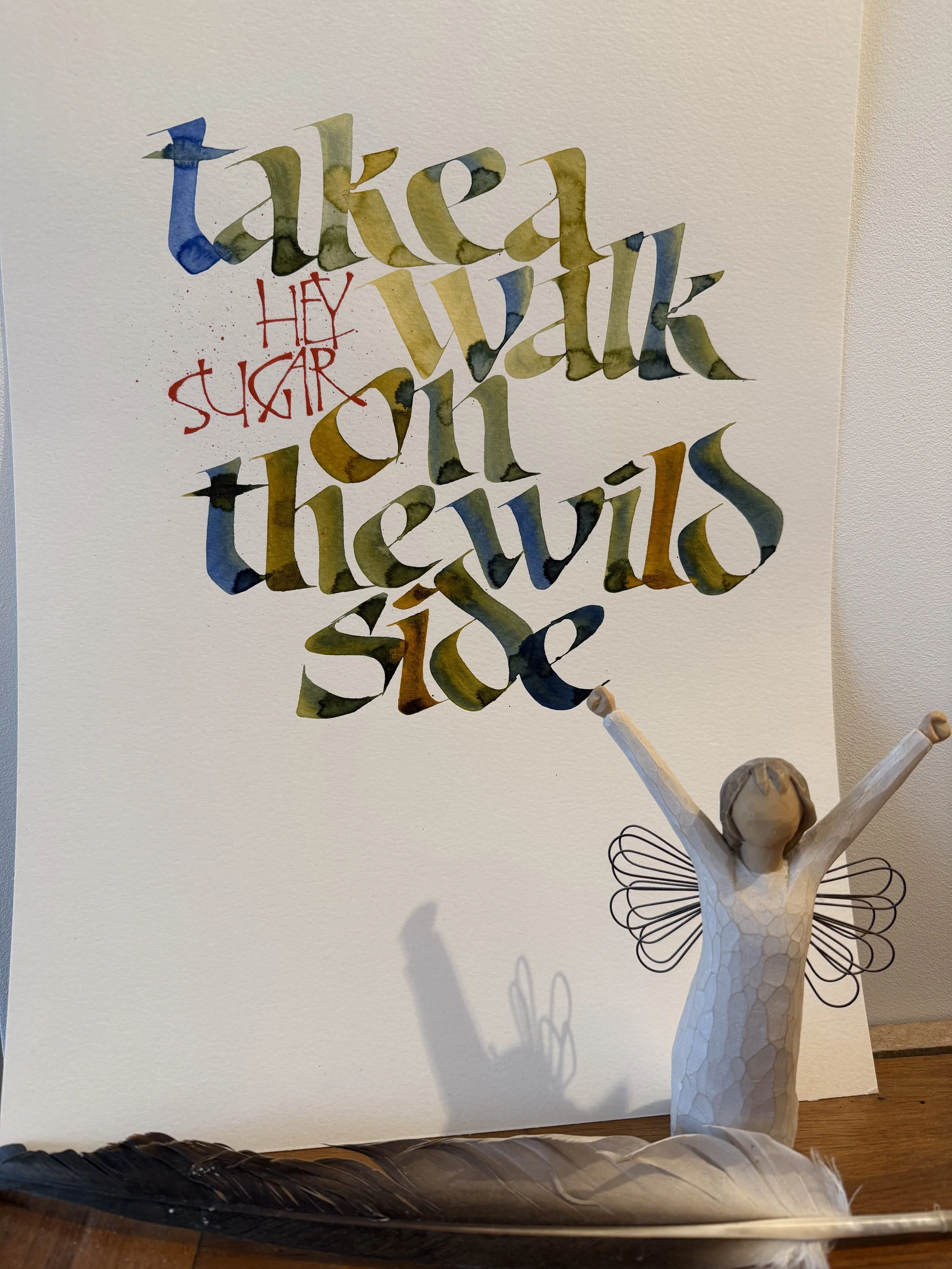 Hand-lettered calligraphy artwork reading “take a walk on the wild side” in watercolor green and blue tones, with “hey sugar” in red script, displayed beside a small angel figurine and a feather